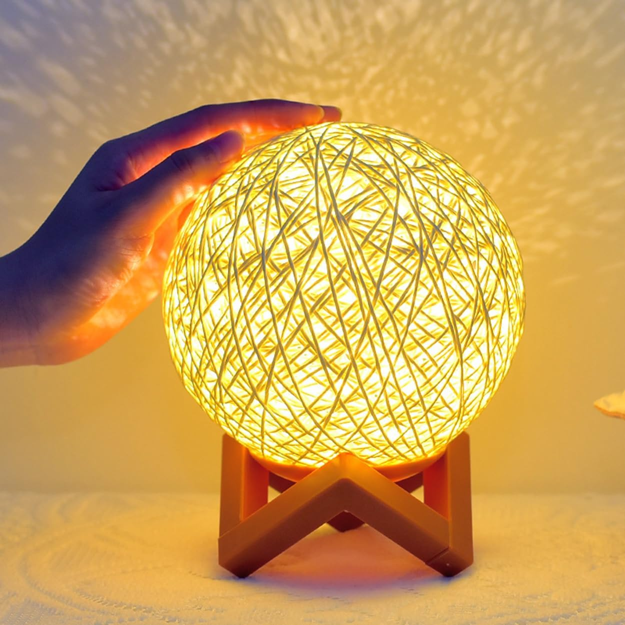 Eine Hand berührt eine leuchtende Rattankugellampe auf einem Holzständer, die ein warmes Licht wirft.