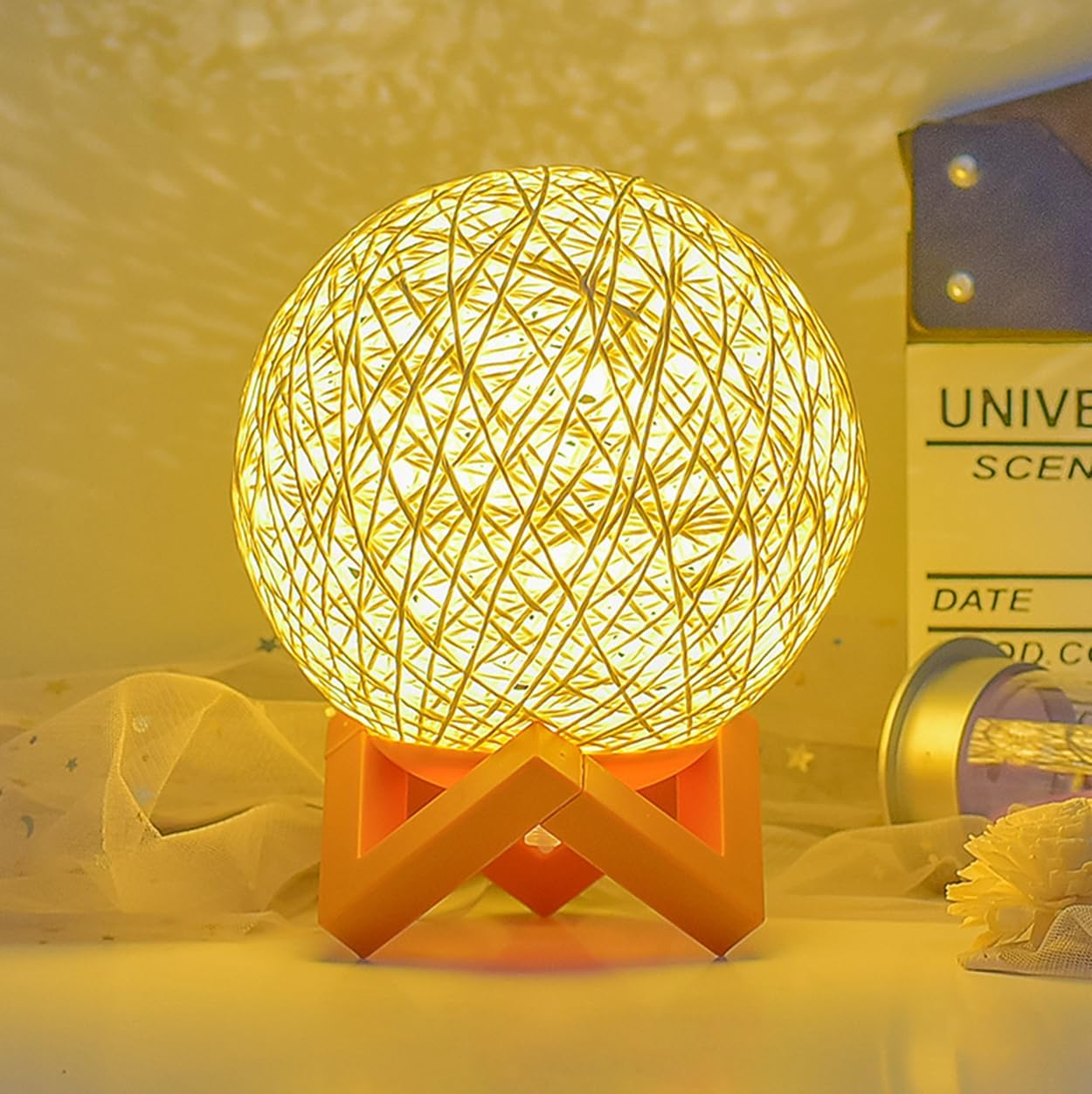 Leuchtende Fadenkugel-Lampe auf orangefarbenem Holzständer, die ein weiches, warmes Lichtmuster an die Wand wirft.