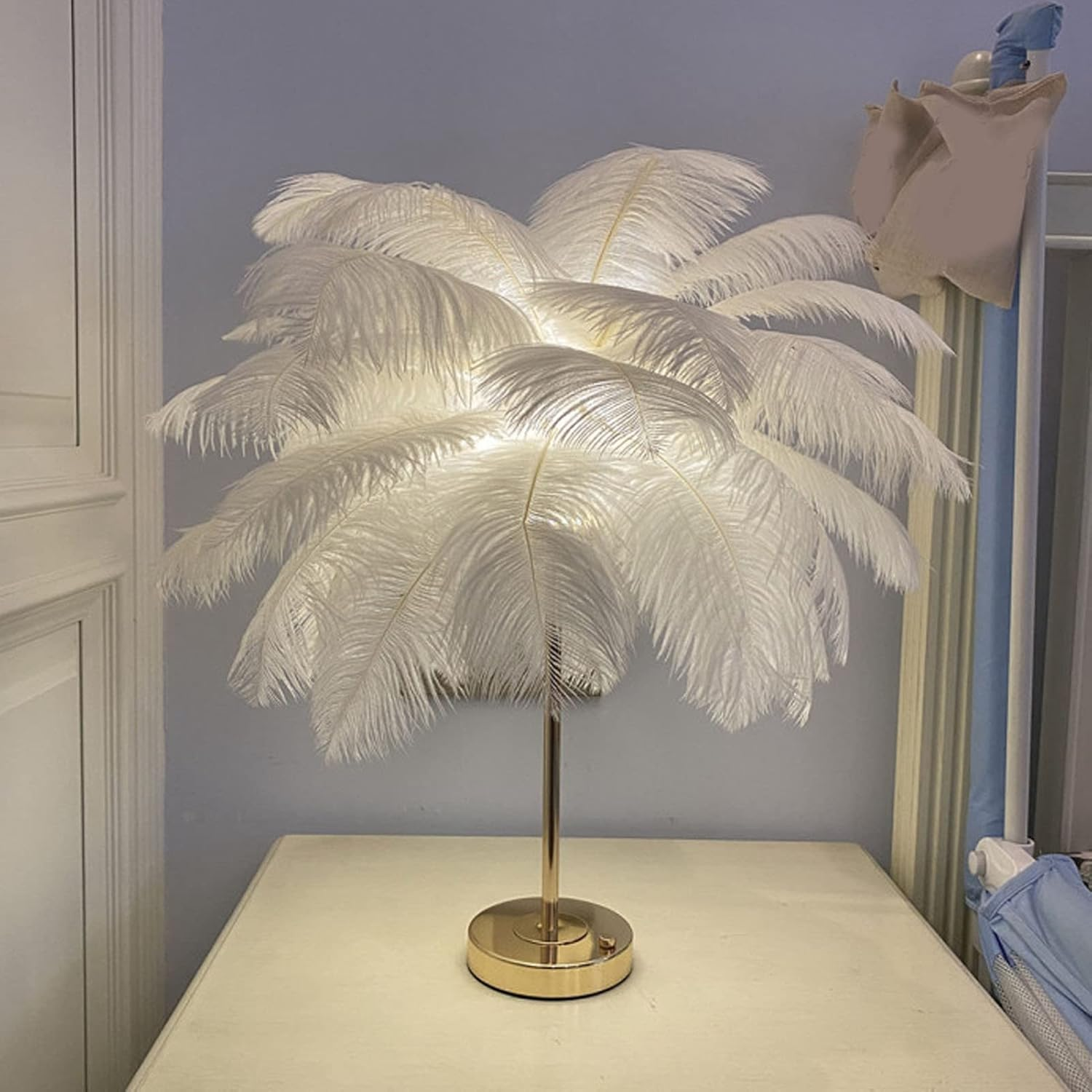 Eine Tischlampe mit weißen Federn und einem goldenen Ständer steht auf einem hellen Tisch.