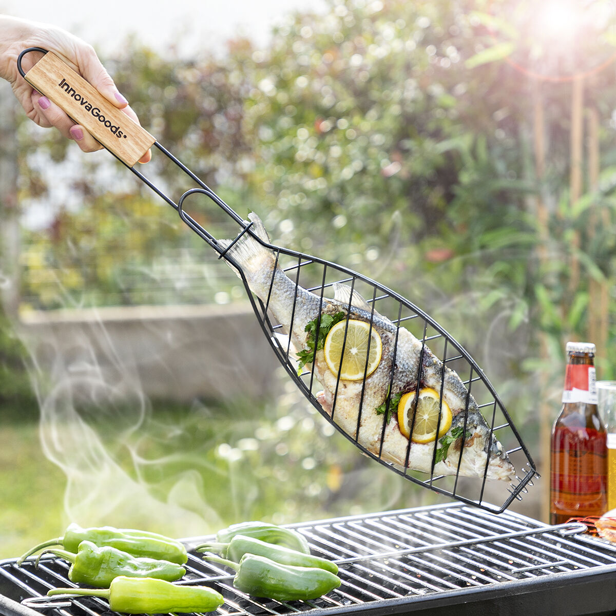 Eine Hand hält einen gegrillten Fisch in einem Drahtkorb über einen Grill mit grünen Paprika und Rauch.