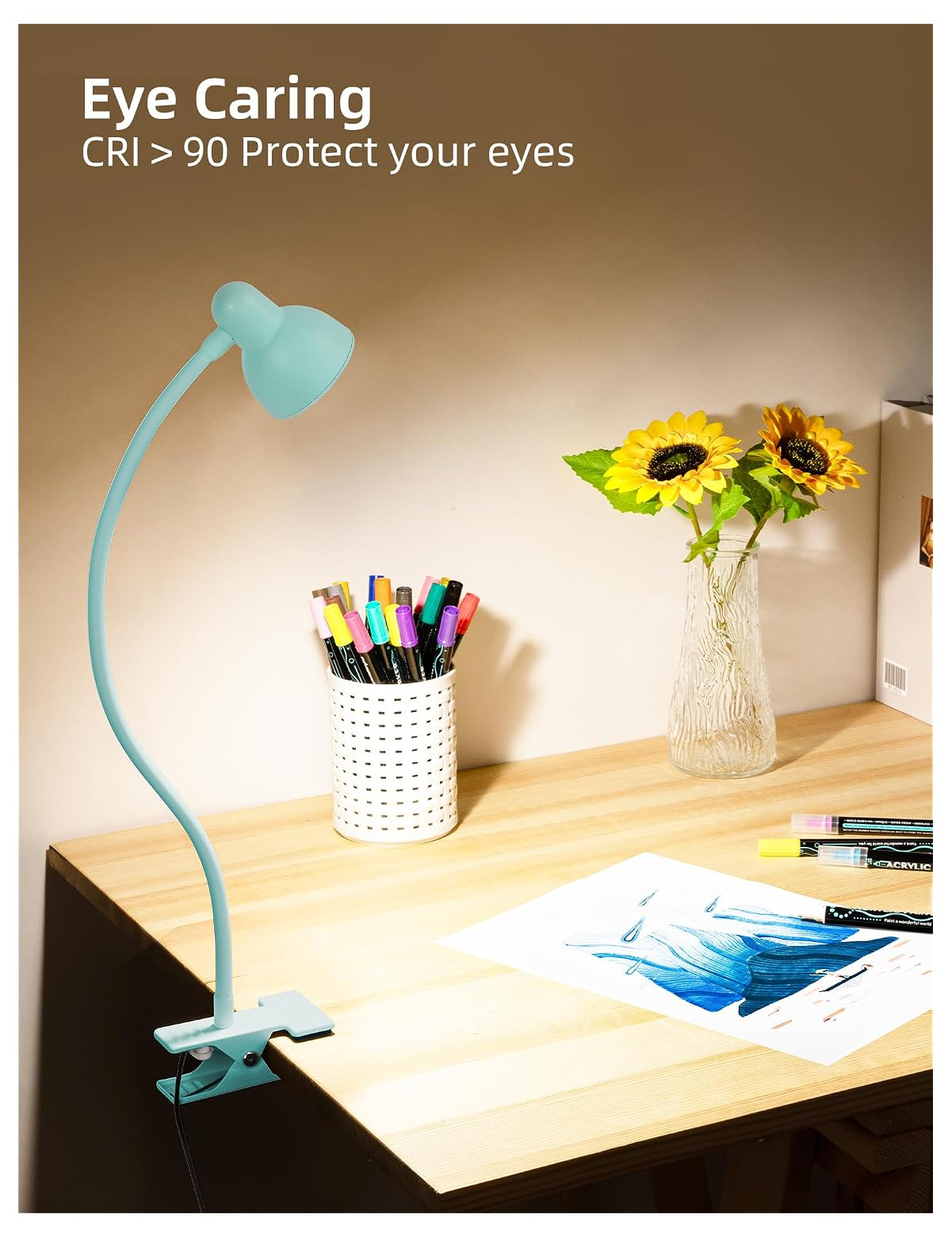 Eine hellblaue Klemmtischlampe beleuchtet einen Holztisch mit Stiftehalter, Sonnenblumen-Vase und blauer Zeichnung.