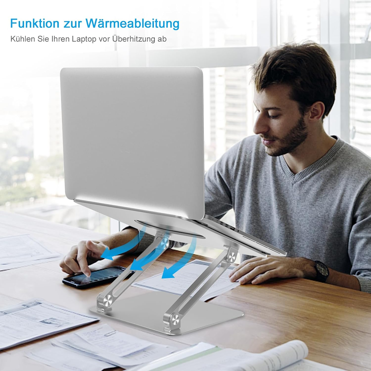 Ein Mann sitzt an einem Holztisch und nutzt einen Laptop auf einem verstellbaren silbernen Metallständer.