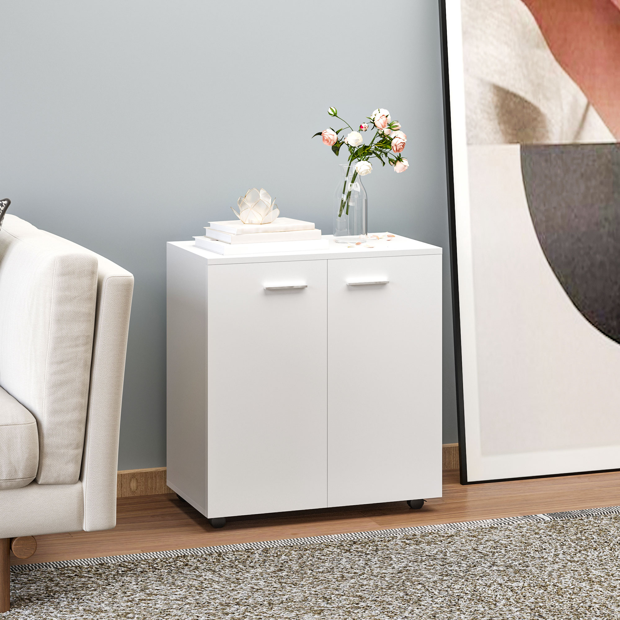 Un mobile bianco con due ante su pavimento in legno, con libri e vaso, vicino a un divano e a un quadro.