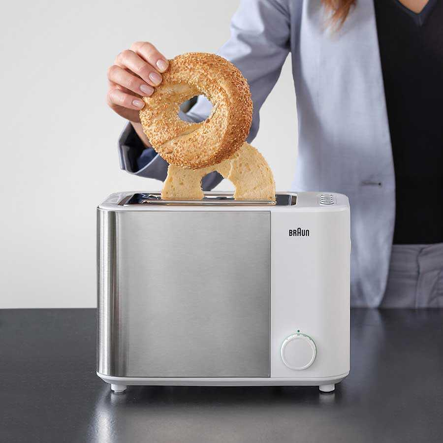 Una persona inserisce un bagel al sesamo tagliato in un moderno tostapane Braun bianco e argento.