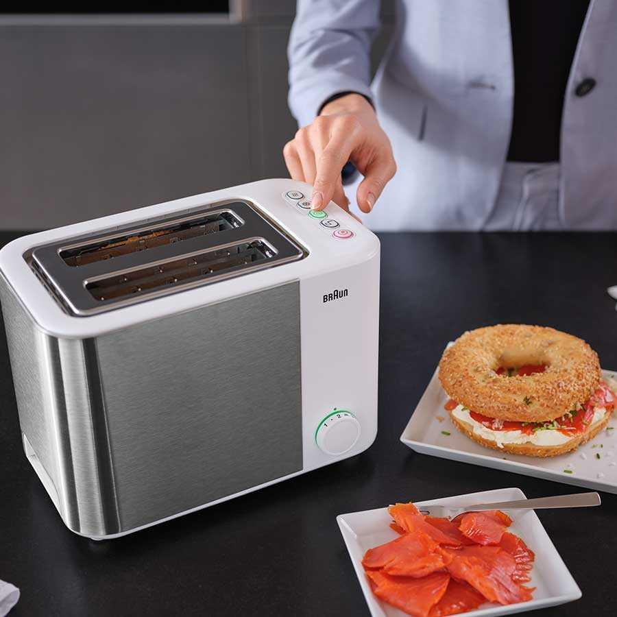 Una persona preme i pulsanti su un tostapane Braun bianco e argento, accanto a piatti con salmone e un bagel.