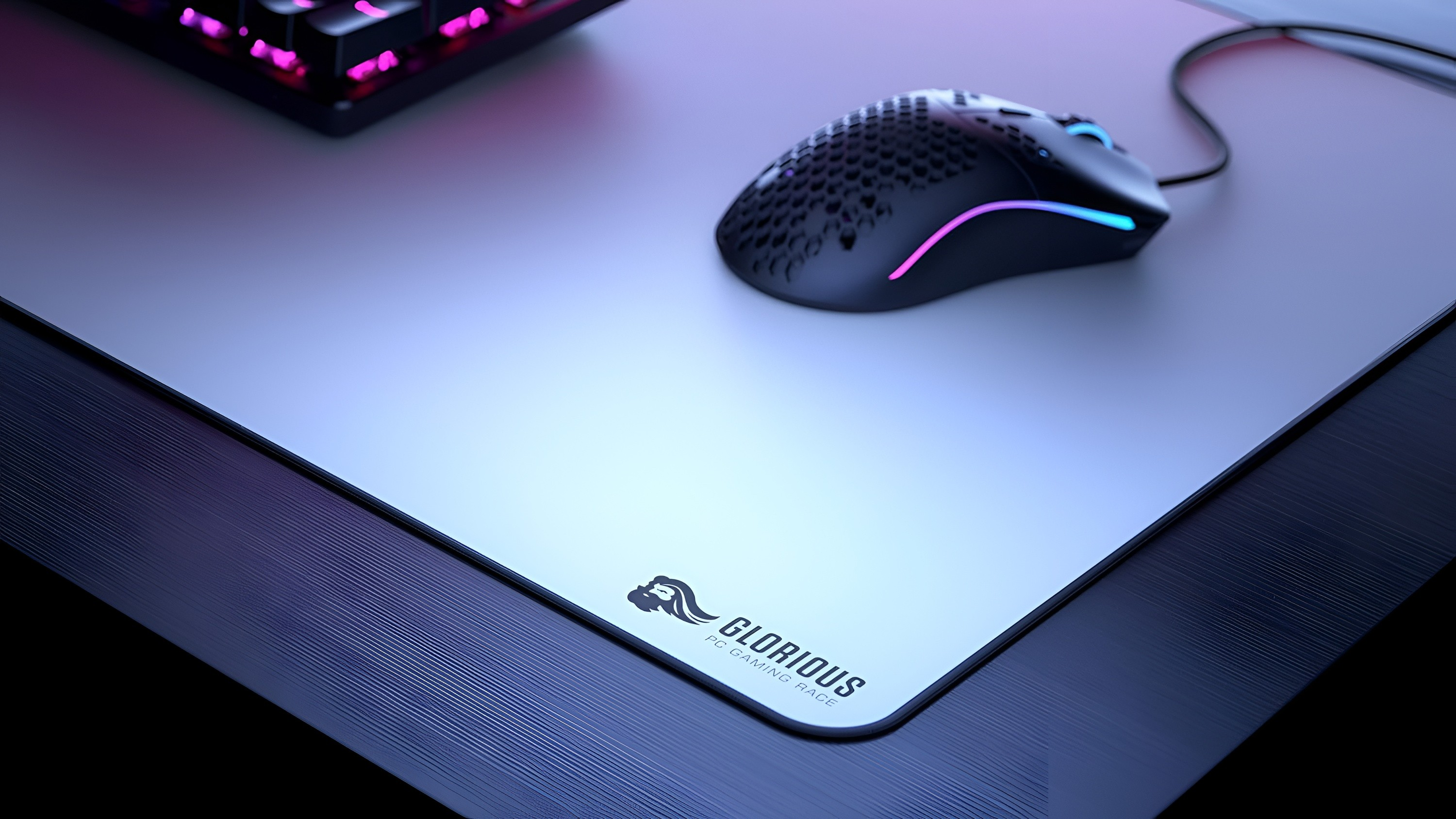 Arka planda bir klavye ile açık renkli Glorious fare altlığı üzerinde RGB aydınlatmalı siyah bir oyuncu fares.