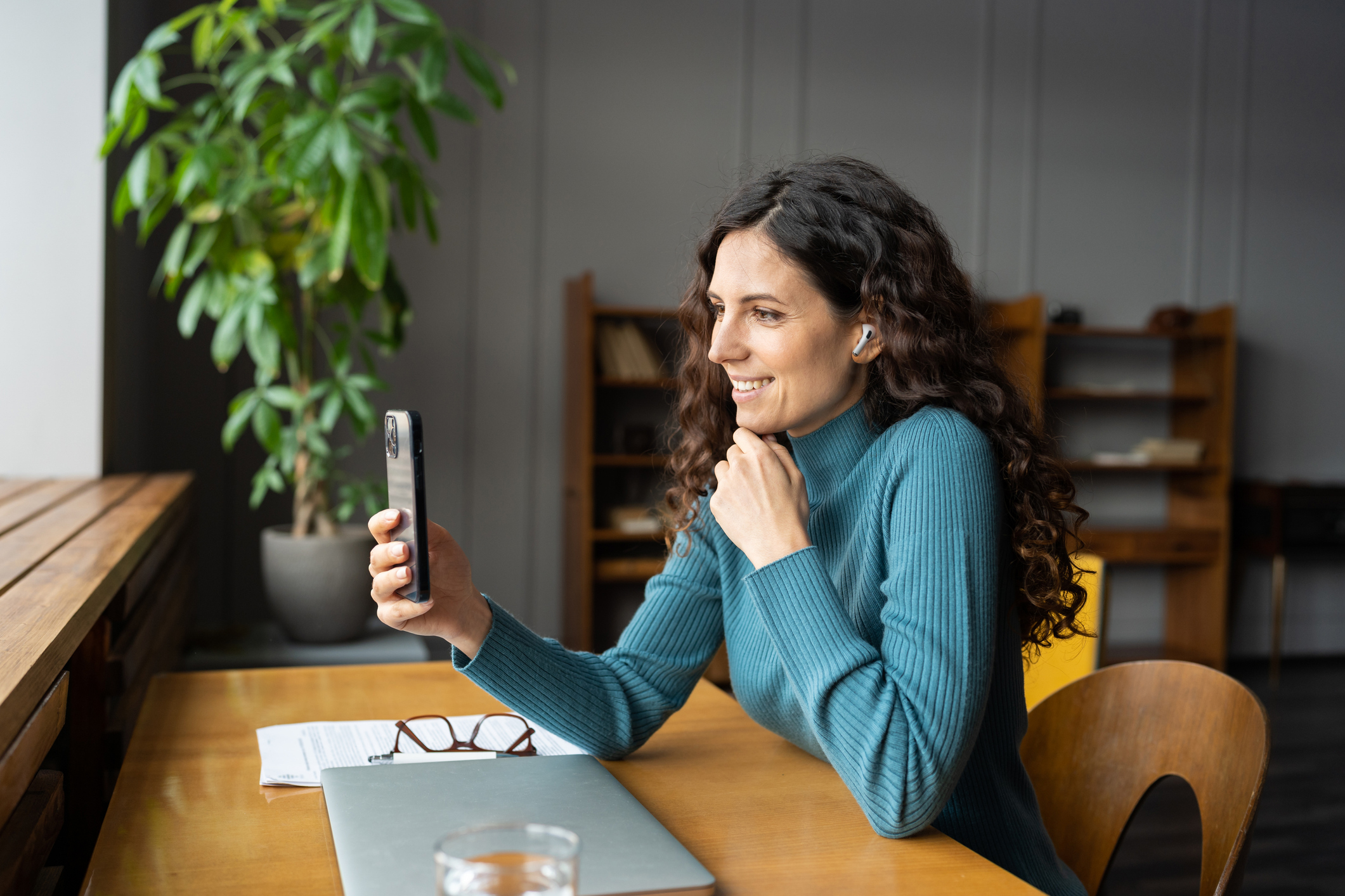 Eine lockere Frau mit dunklen Locken und blauem Pullover sitzt im Café und macht ein Selfie mit dem Smartphone.