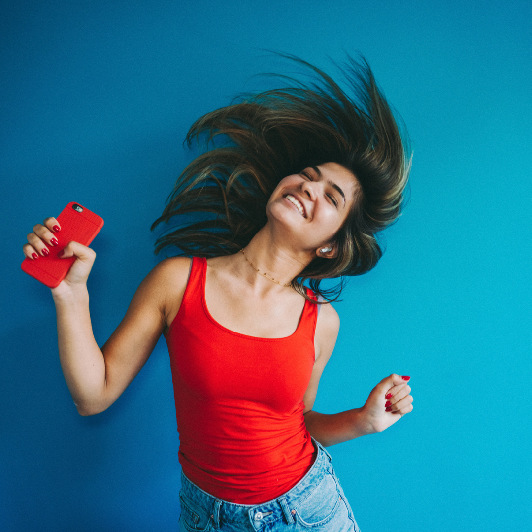 Eine lächelnde Frau in rotem Tanktop wirft ihr braunes Haar vor einem blauen Hintergrund zurück und hält ein rotes Smartphone.