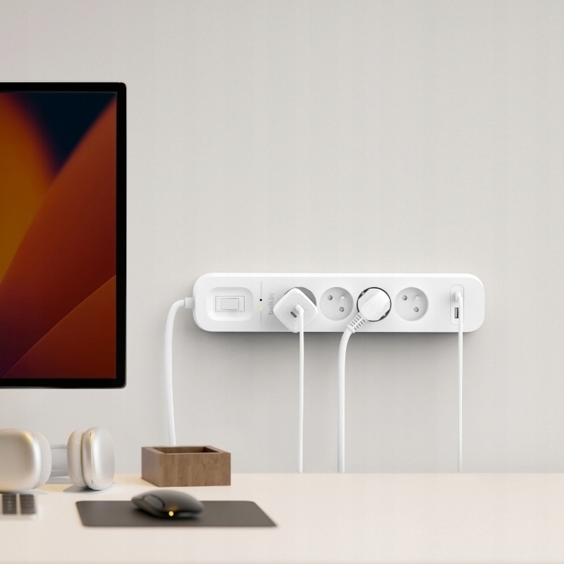 Una ciabatta bianca Belkin montata a parete con cavi collegati, vicino a un monitor e accessori da scrivania.