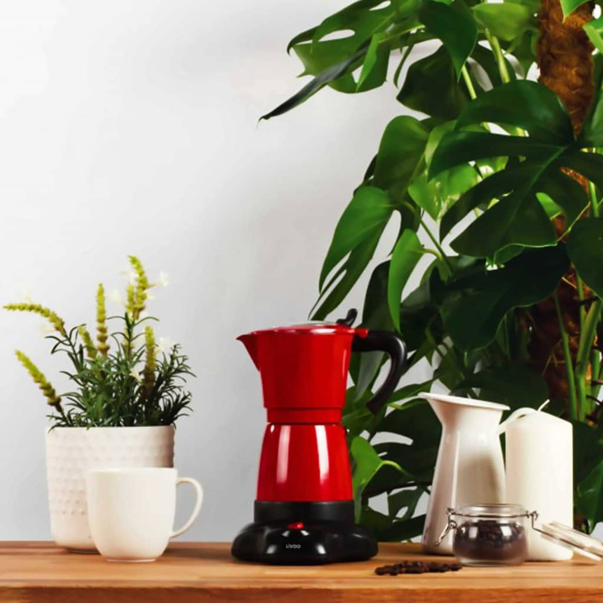 Eine rote elektrische Mokkakanne auf einem Holztisch mit grünen Pflanzen, einer weißen Tasse und einem weißen Krug.