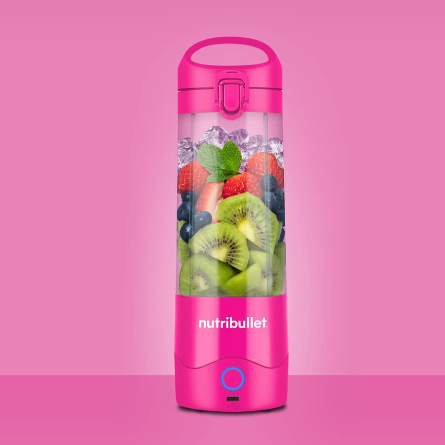 A pink Nutribullet blender filled with strawberries, kiwis, blueberries, and mint against a pink background.
