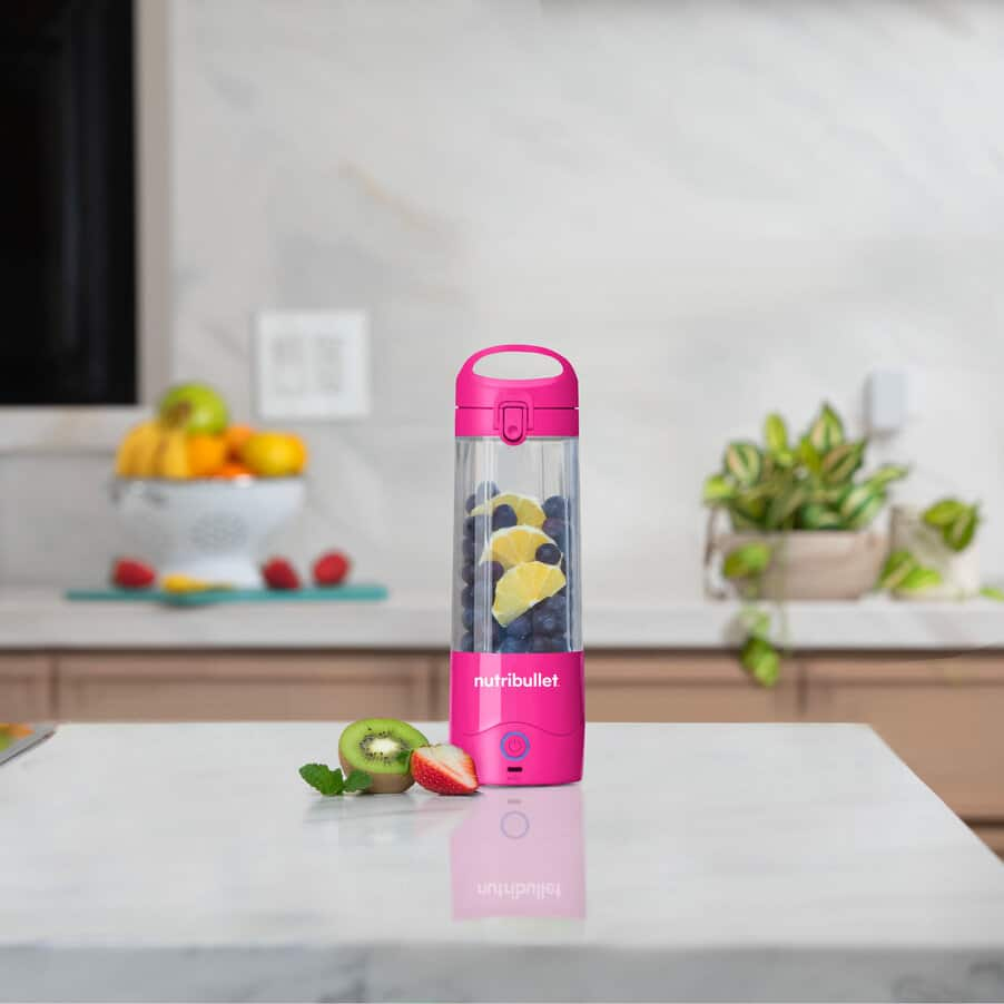 A pink Nutribullet blender filled with blueberries and lemon slices sits on a white counter in a kitchen.