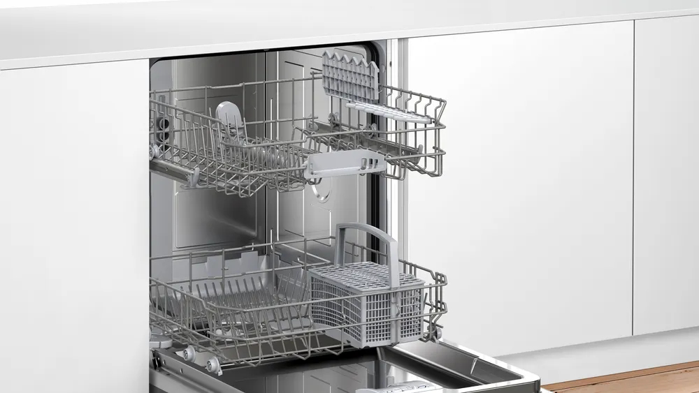 Lavastoviglie bianca aperta con cestelli grigi estraibili vuoti e un cestello portaposate in una cucina moderna.