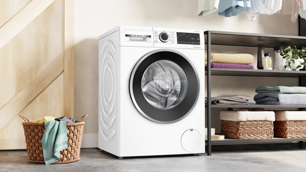 Eine weiße Bosch-Waschmaschine steht in einem hellen Waschraum neben einem Regal mit Handtüchern und einem Korb.