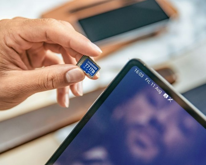 Una mano tiene una minuscola scheda di memoria Samsung T7 da 1TB vicino a un tablet.