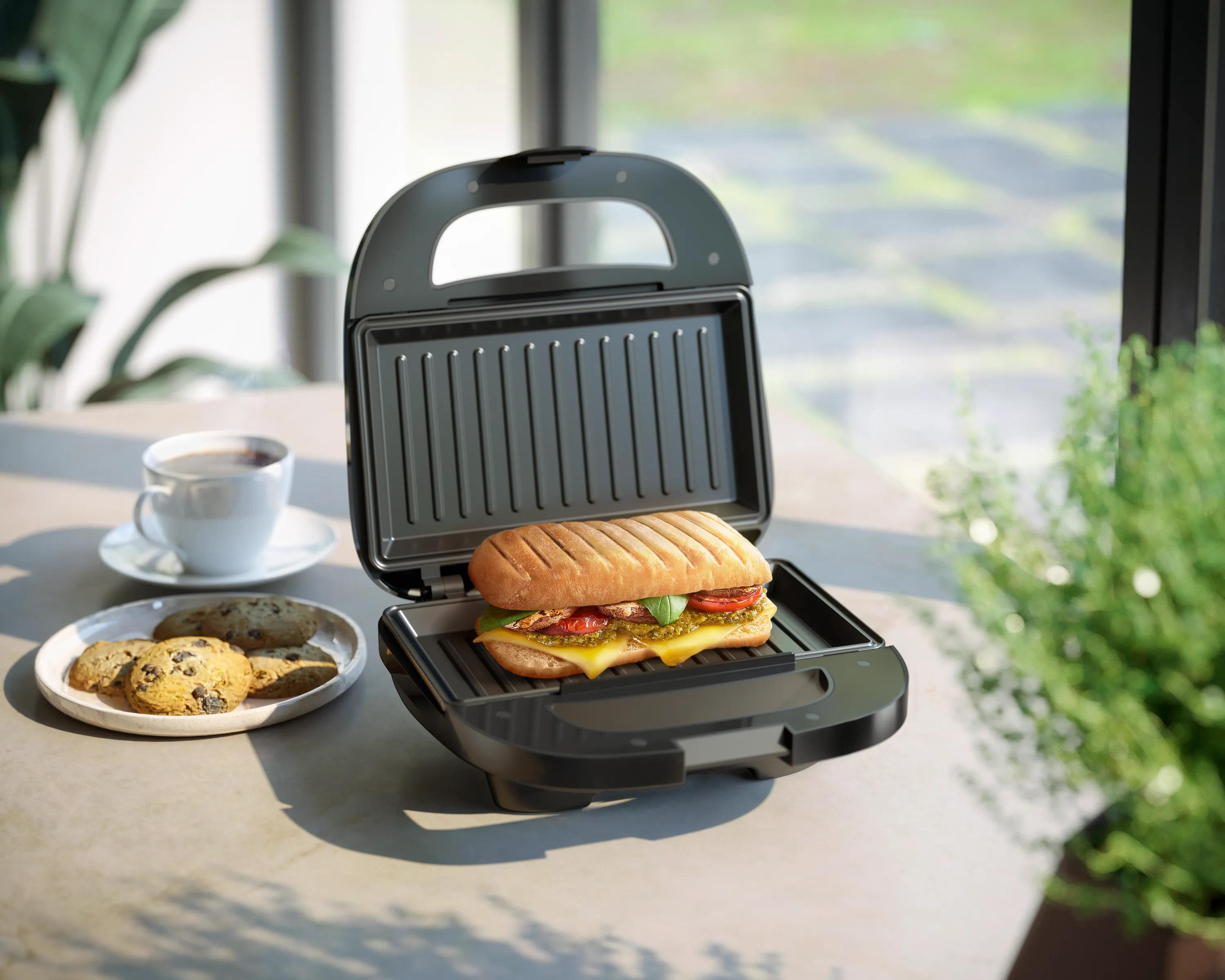 Otwarty opiekacz do kanapek na stole z panini w środku, obok ciasteczek i filiżanki kawy.