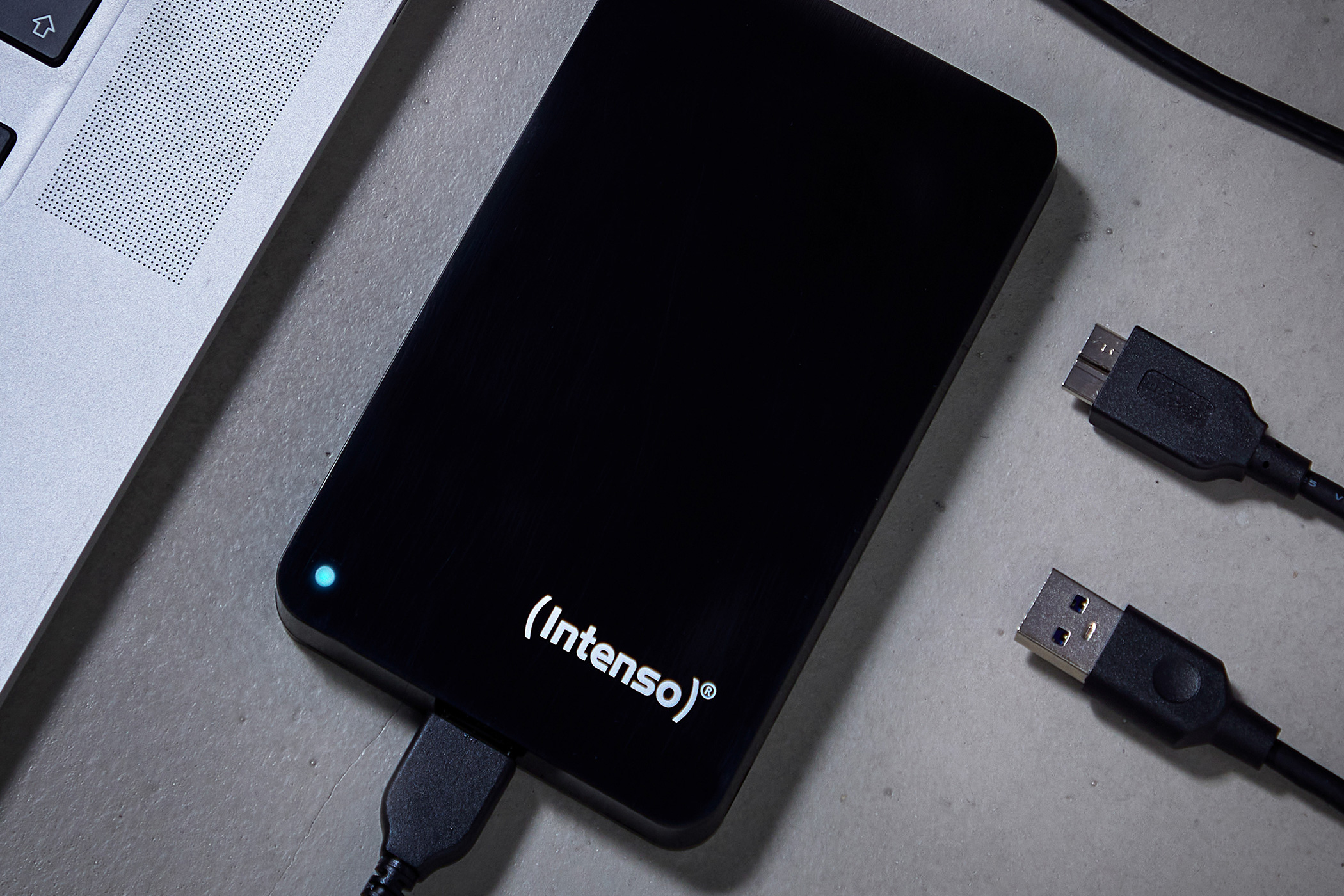 Eine schwarze Intenso-Festplatte mit leuchtend blauer LED liegt neben einem Laptop und USB-Kabeln.