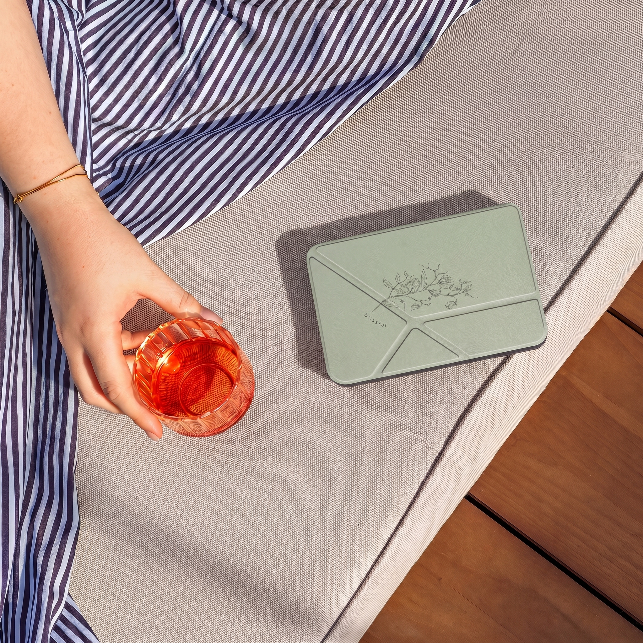 Een persoon in een gestreepte jurk houdt een roodachtige drank vast naast een groen etui op een ligstoel.