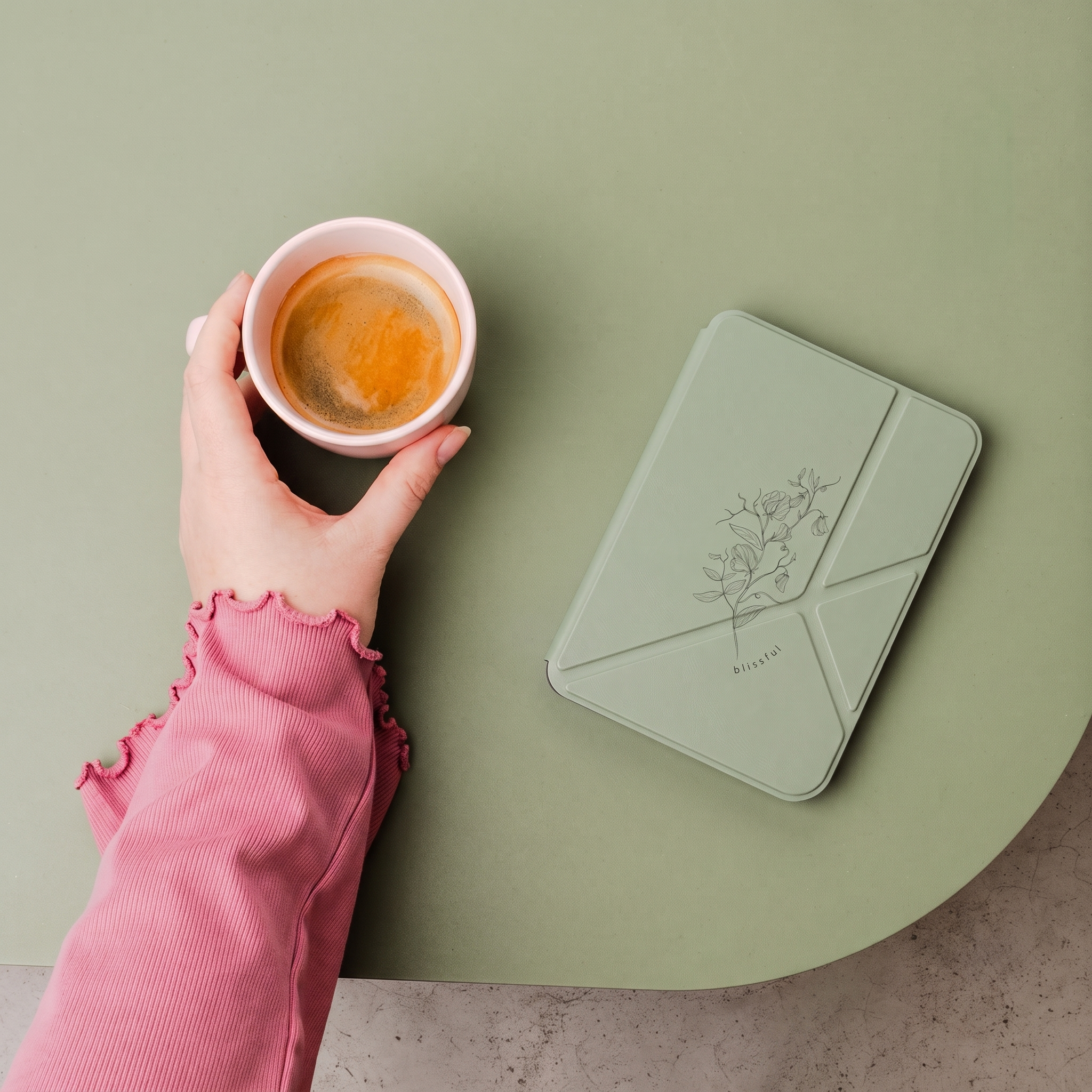 Een hand in een roze trui houdt een kop koffie vast naast een groen notitieboek op een groene tafel.