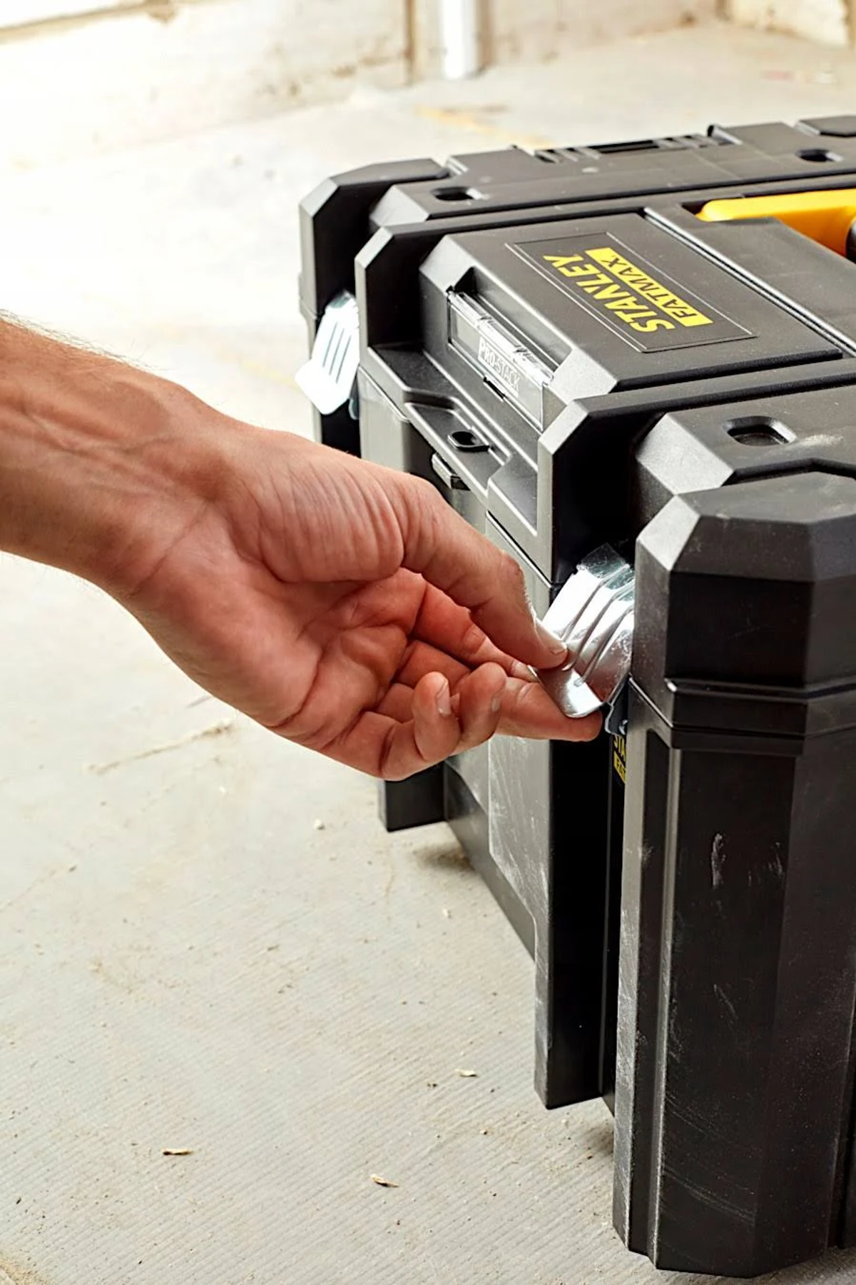 Eine Hand öffnet die Metallverriegelung eines schwarzen Stanley FatMax Werkzeugkoffers auf dem Boden.