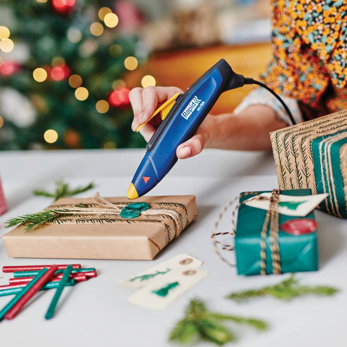 Eine Hand benutzt eine blaue Heißklebepistole, um grüne Siegelwachs auf ein Weihnachtsgeschenk aufzutragen.