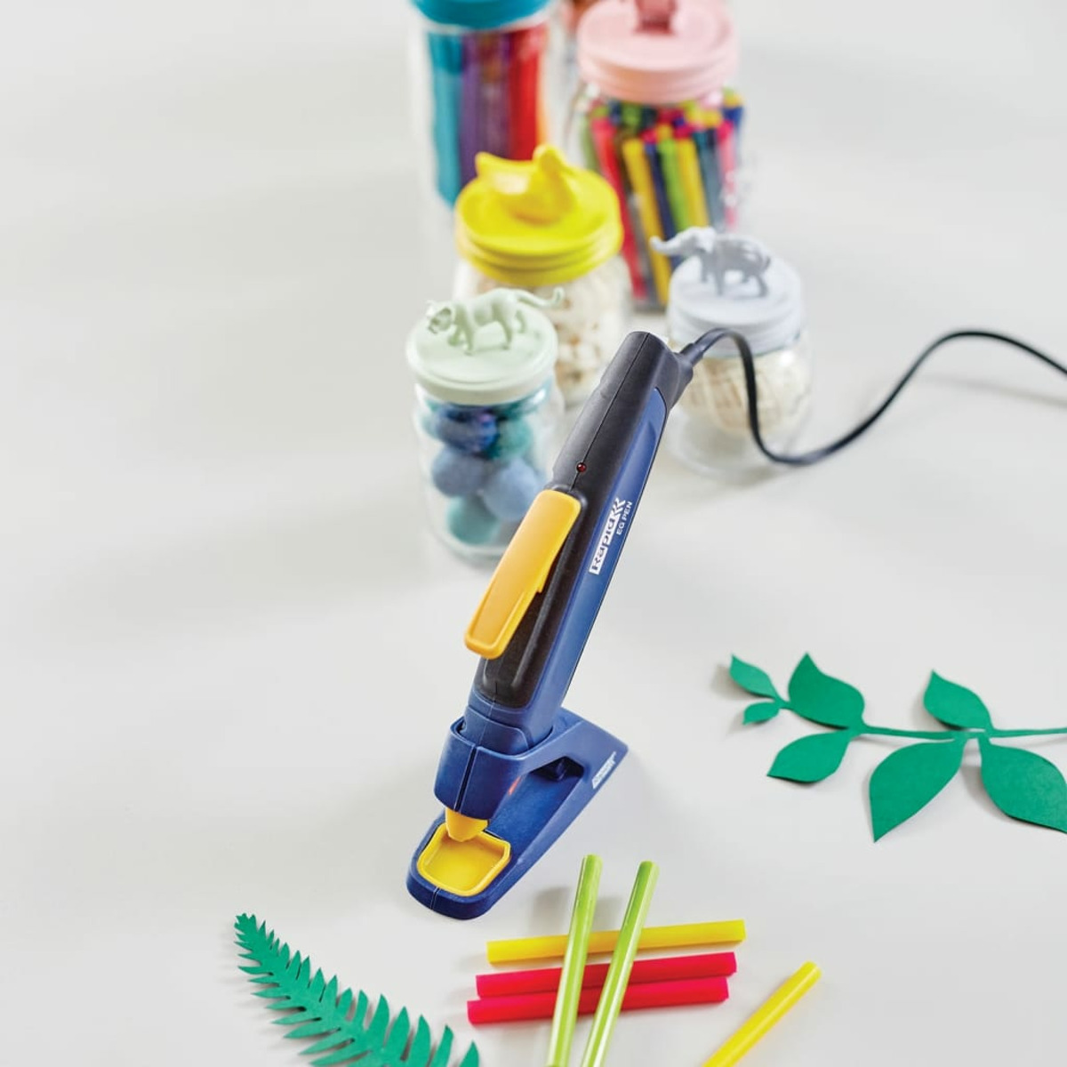 Blaue Heißklebepistole auf weißem Tisch, umgeben von bunten Klebestiften, Papierblättern und Gläsern.