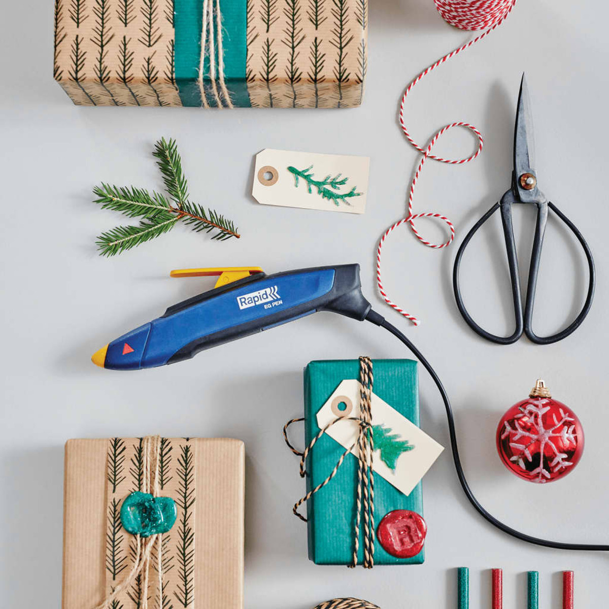 Geschenkverpackungszubehör mit Geschenkpapier, Schere, Heißklebepistole, Tannenzweigen und roten Weihnachtskugeln.