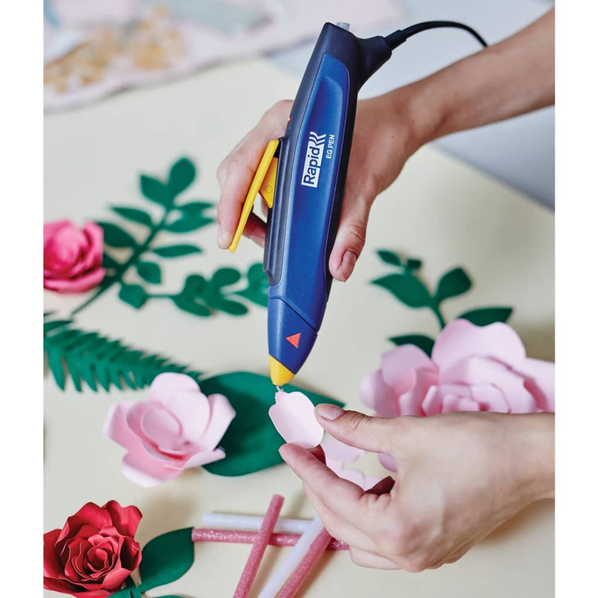 Eine Hand hält eine blaue Heißklebepistole, um rosa Papierblumen und grüne Blätter auf einem Tisch zu basteln.