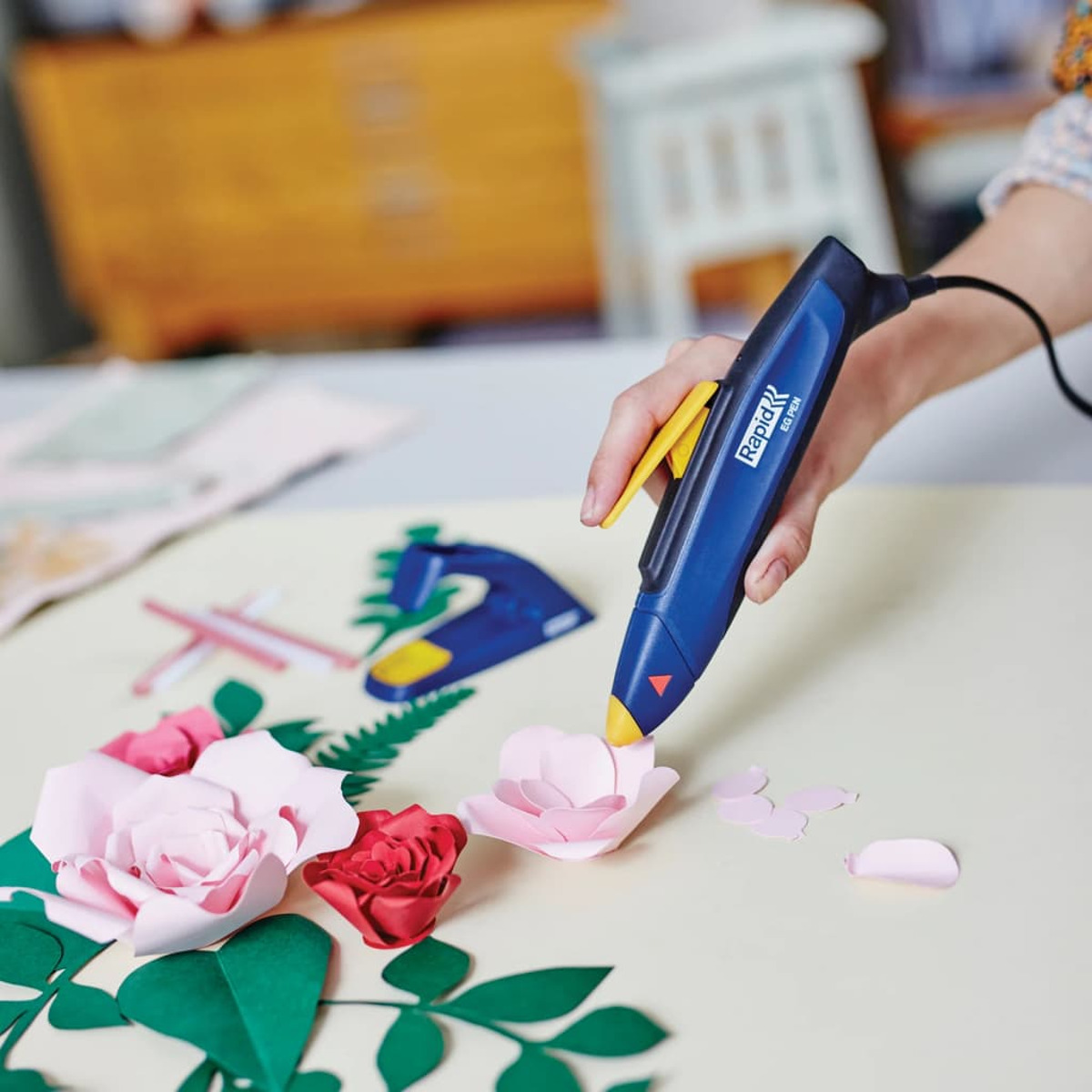 Eine Hand benutzt eine blaue Heißklebepistole, um Papierblumen und Grünelemente auf einer weißen Fläche zusammenzukleben.