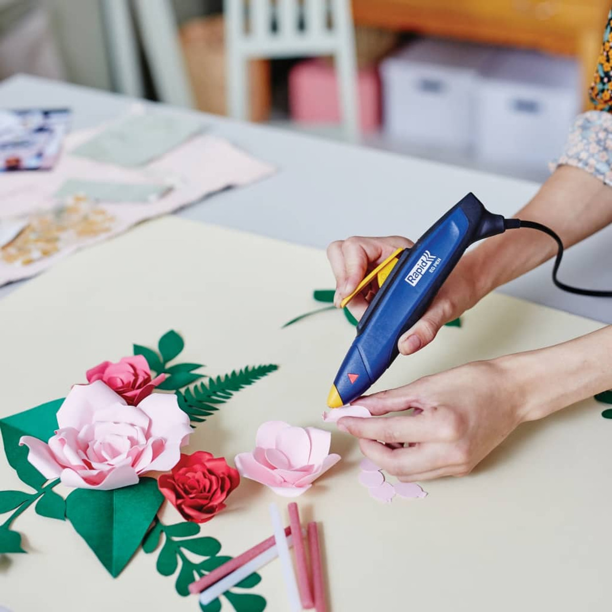 Eine Person verwendet eine blaue Heißklebepistole, um rosa Papierblumen und grüne Blätter auf einem Tisch zusammenzusetzen.