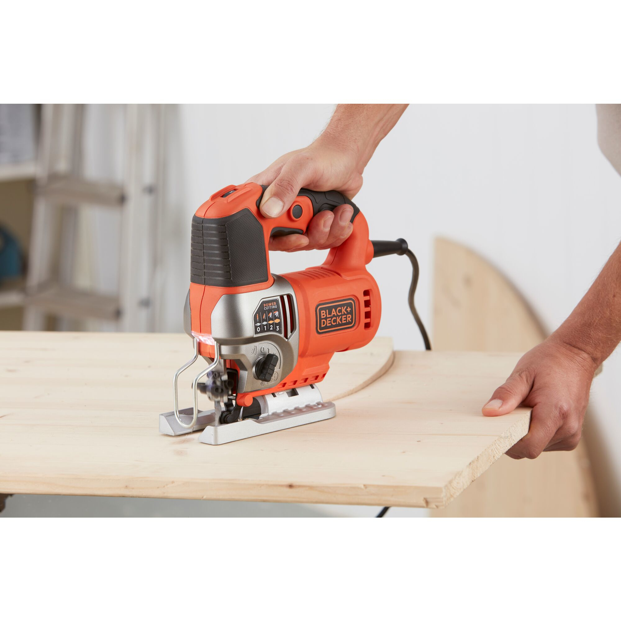 Eine Hand hält eine orange-schwarze Stichsäge und schneidet eine Holzplatte in einer Werkstatt.