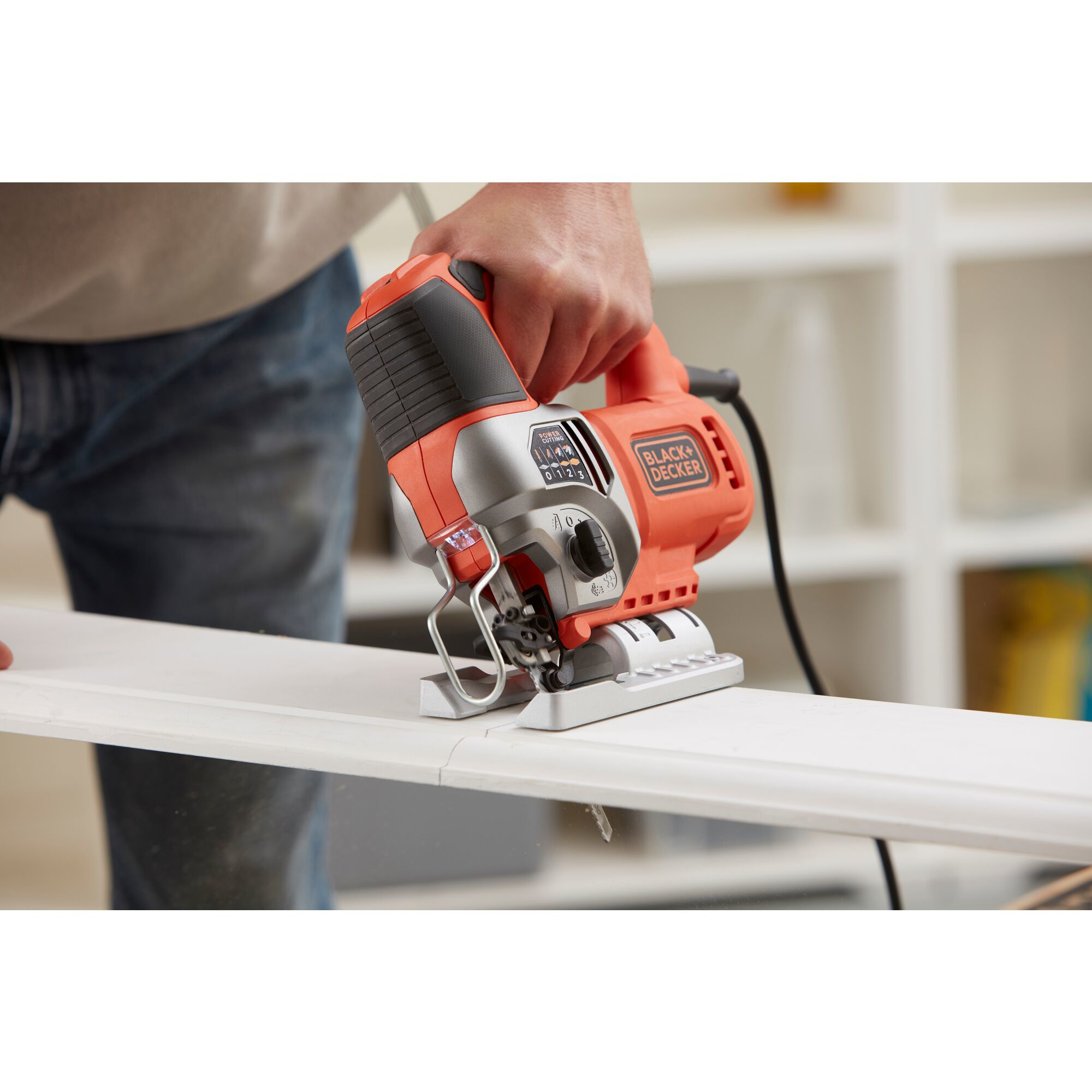 Eine Person schneidet ein weißes Holzbrett mit einer orange-schwarzen Black+Decker Stichsäge in einer Werkstatt.