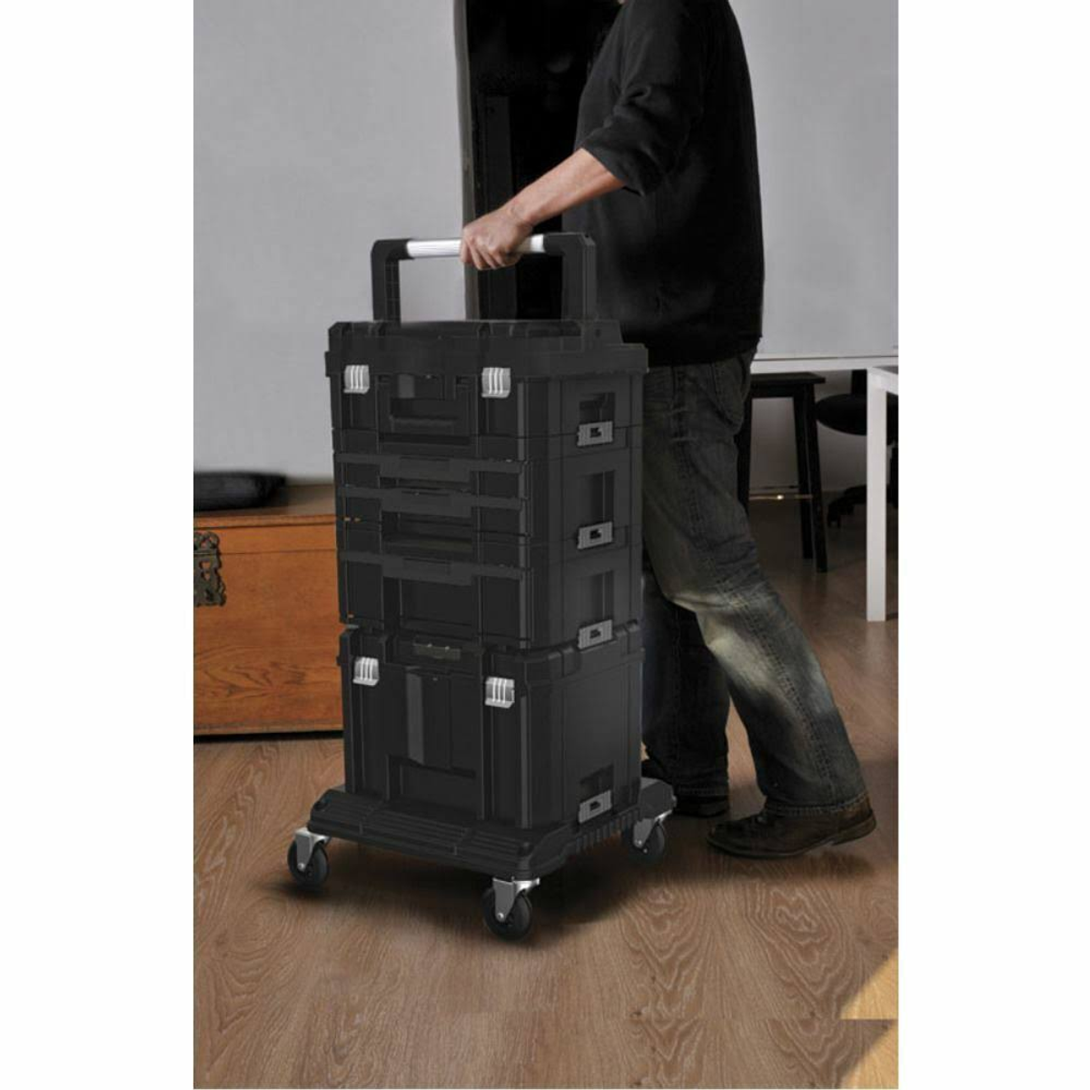 Eine Person zieht eine gestapelte, schwarze Werkzeugbox mit Rollen über einen Holzboden.