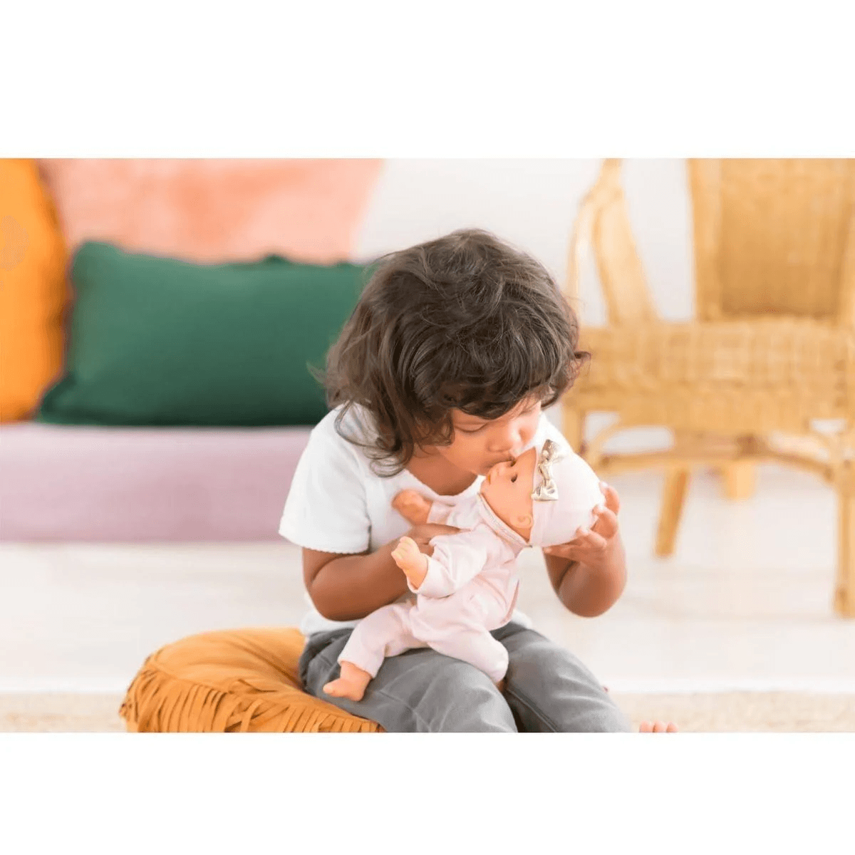 Ein kleines Kind mit dunklen Locken sitzt auf einem Kissen und küsst zärtlich eine Puppe.