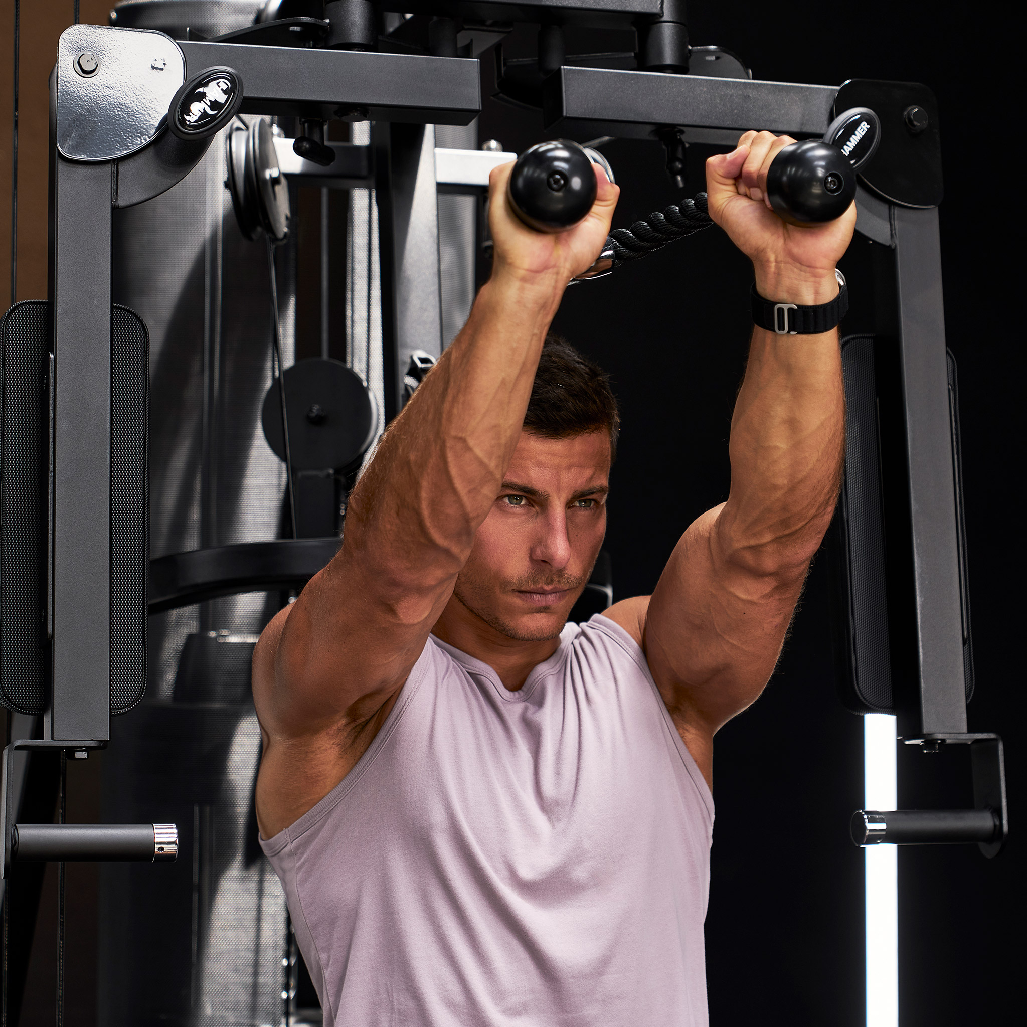 Ein muskulöser Mann in einem grauen Tanktop trainiert an einer schwarzen Seilzugmaschine im Fitnessstudio.