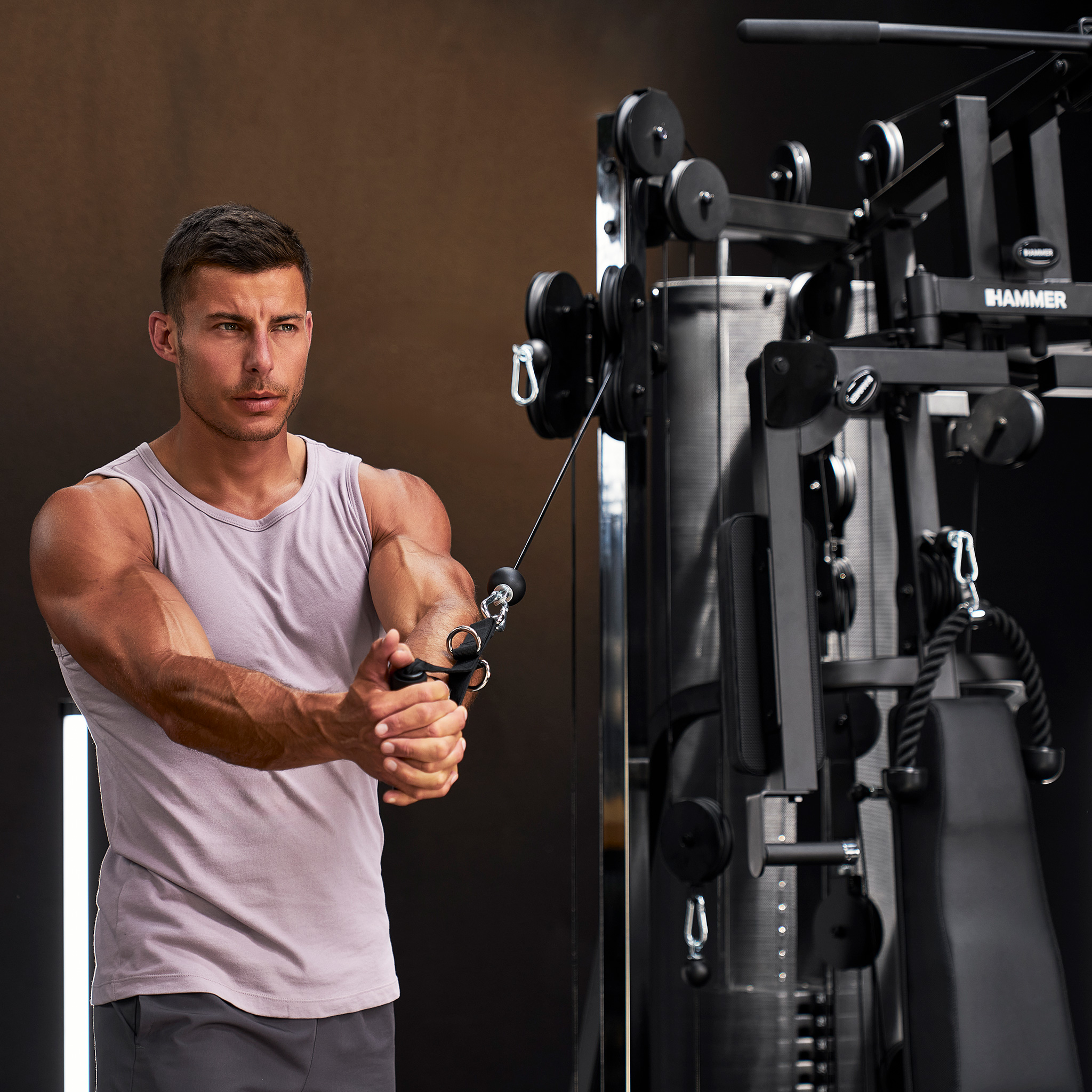 Muskulöser Mann in grauem Tanktop trainiert an einer schwarzen Seilzugmaschine im Fitnessstudio.