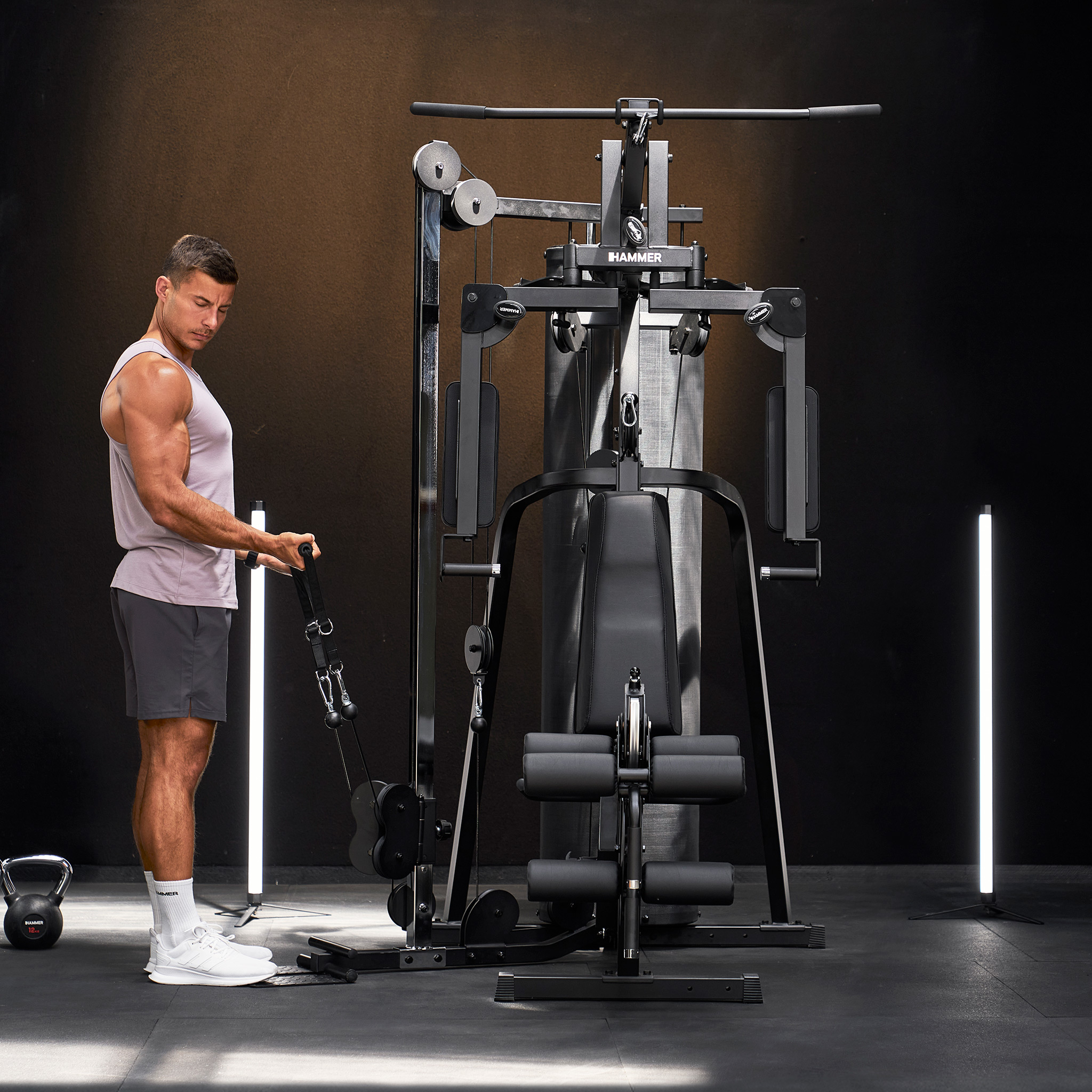 Ein Mann trainiert im Fitnessstudio an einer schwarzen Kraftstation vor dunkelbraunem Hintergrund mit zwei Lichtröhren.