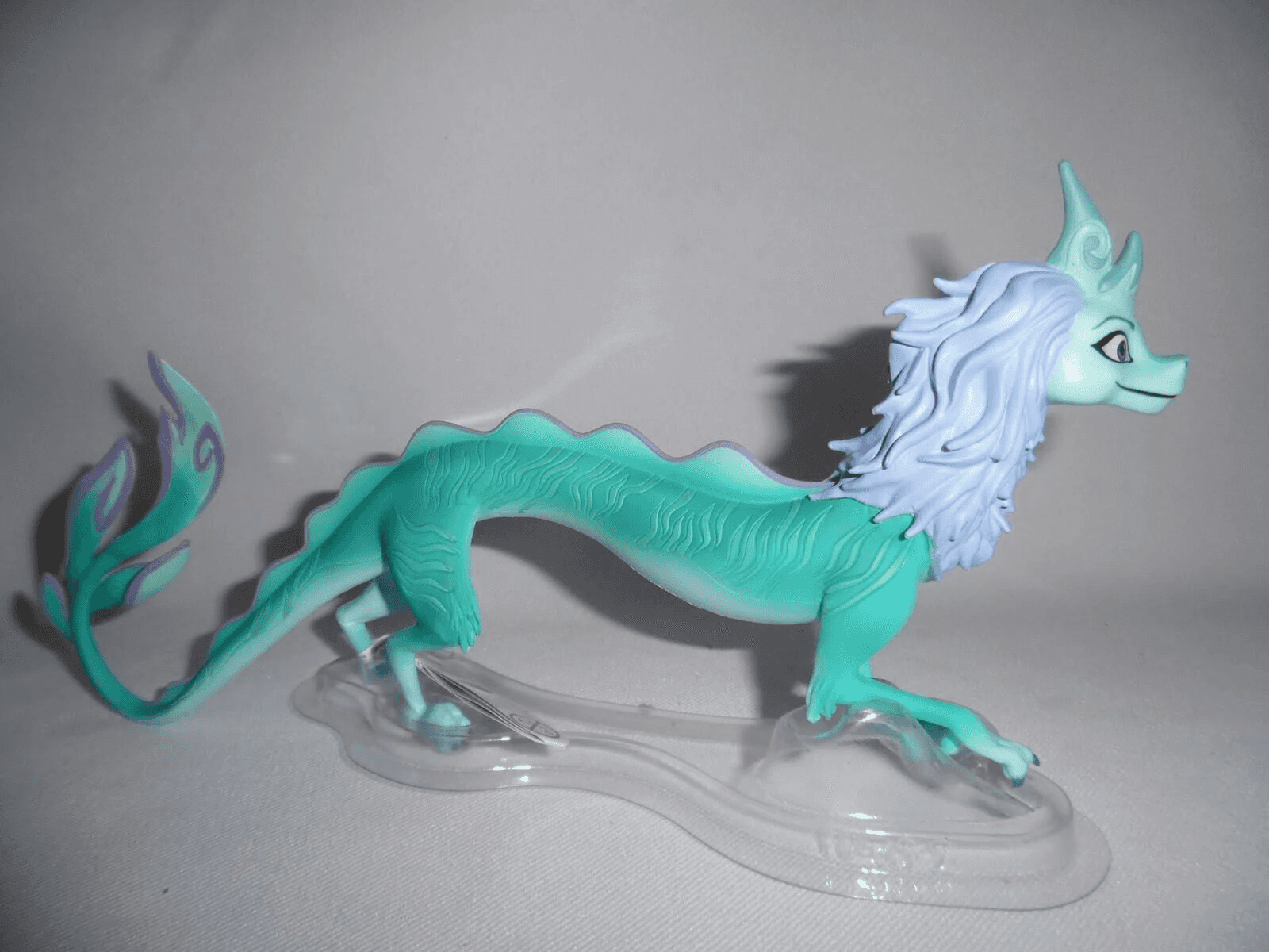 Eine türkisfarbene und lila Drachenfigur mit langer weißer Mähne steht auf einem klaren Kunststoffsockel vor einfarbigem Hintergrund.