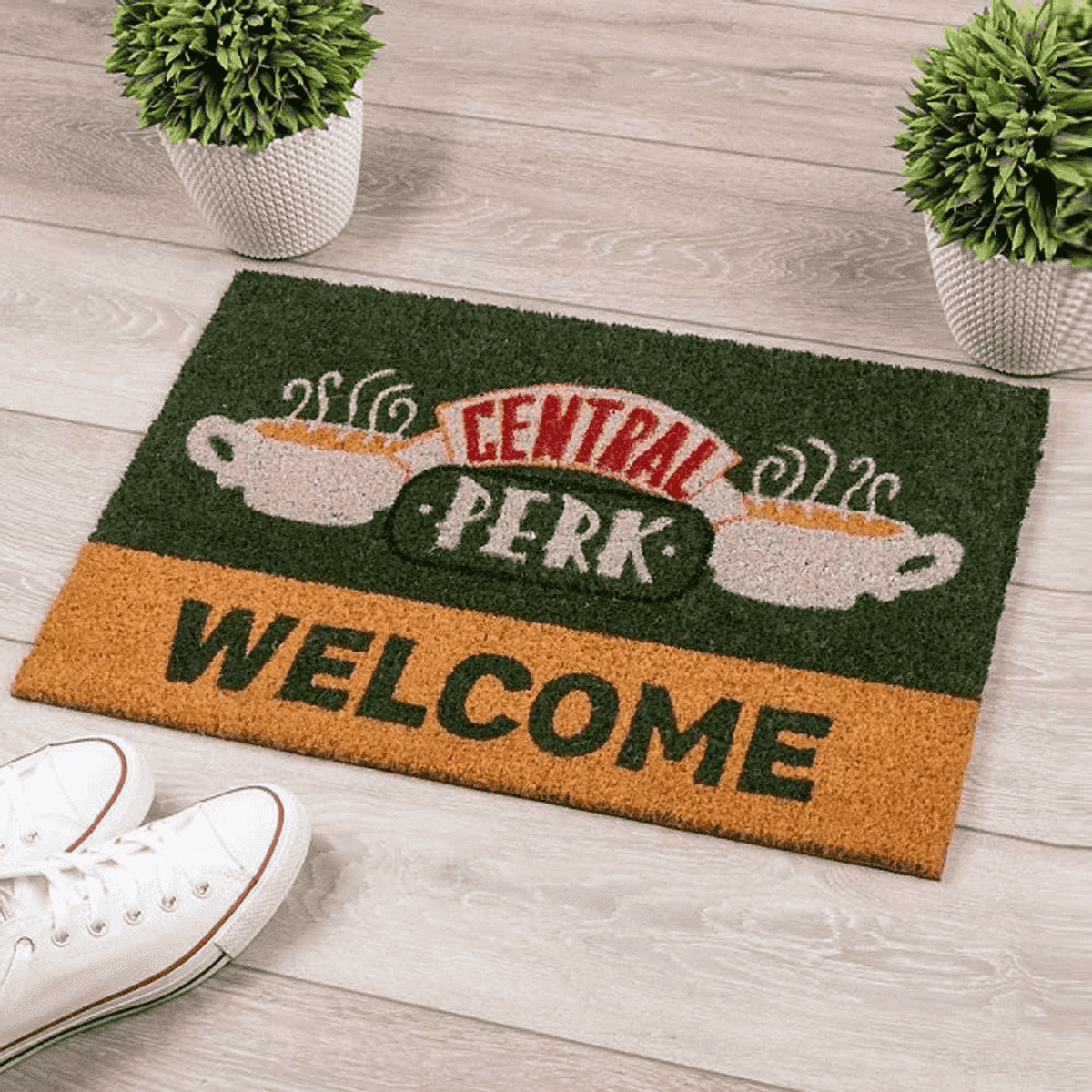 Un felpudo con texto Central Perk y Welcome en suelo de madera, flanqueado por dos plantas y zapatillas blancas.
