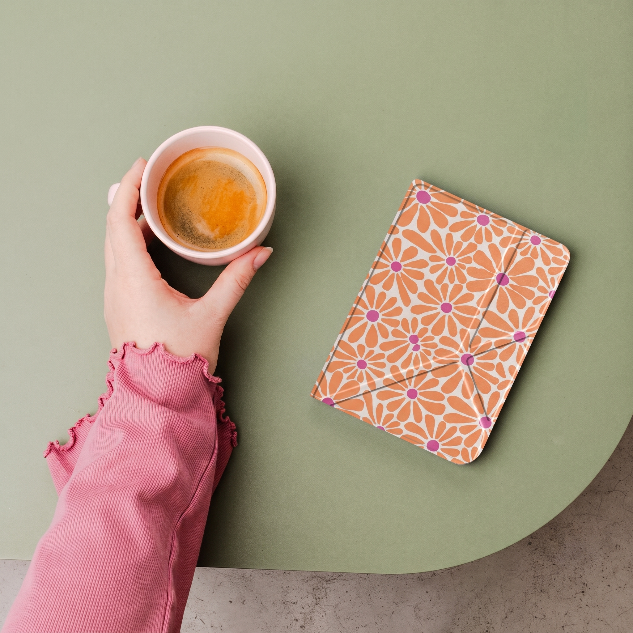 Une main dans un pull rose tient une tasse de café à côté d'un livre fleuri orange sur une table verte.