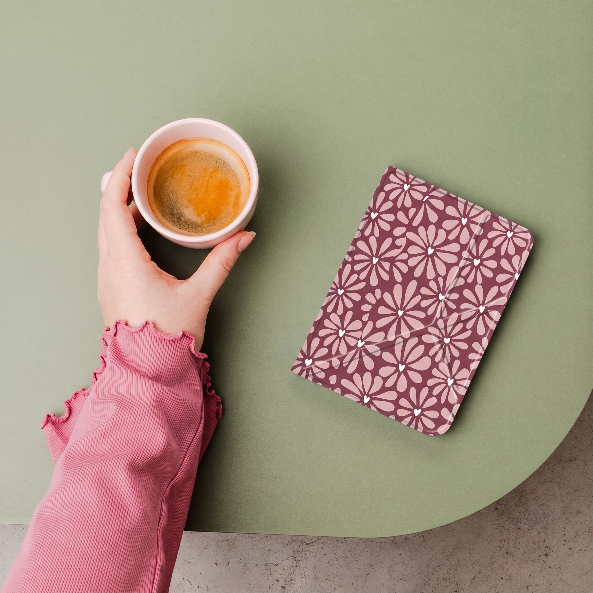 Een hand in een roze trui houdt een koffiekopje vast naast een notitieboekje op een groene tafel.