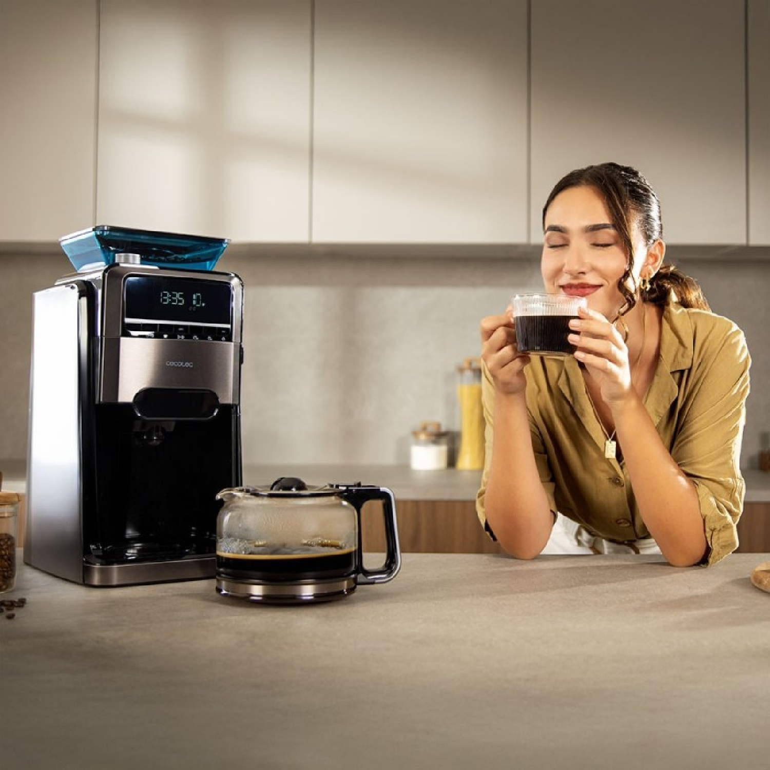 Una donna sorridente tiene una tazza di caffè accanto a una macchina moderna e brocca di vetro.