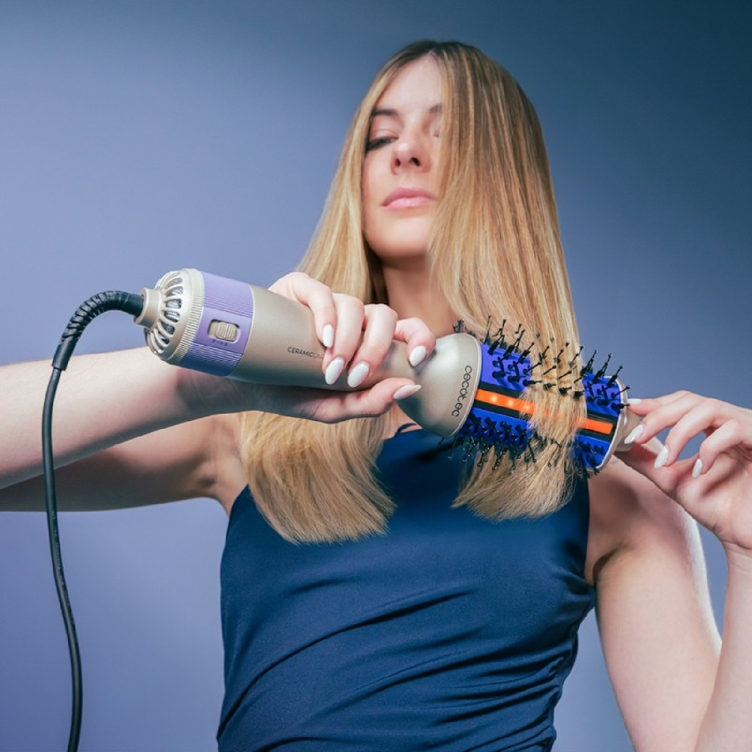 Una donna bionda con un top blu modella i suoi lunghi capelli lisci con una spazzola ad aria calda su sfondo grigio.