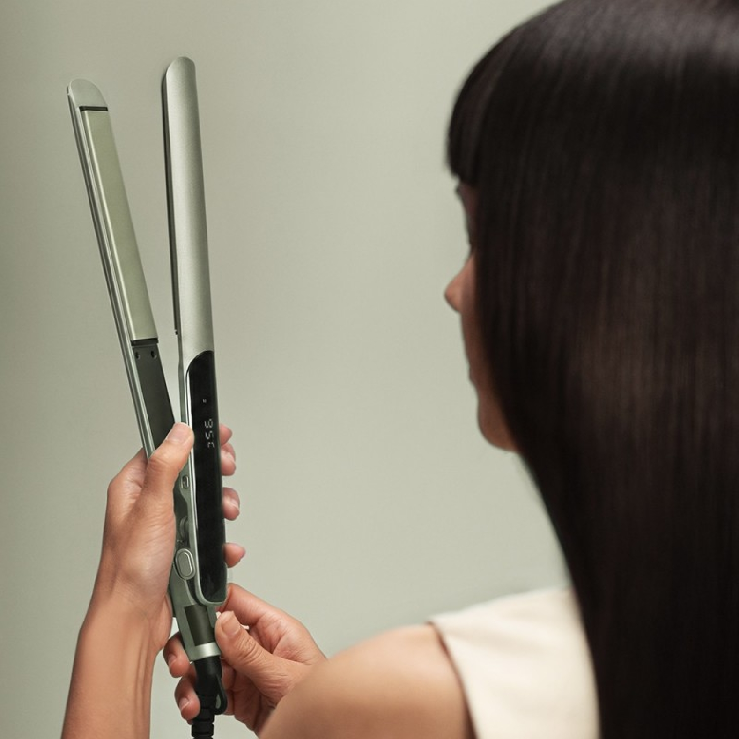 Una donna con capelli neri lisci tiene in mano una piastra per capelli argentata aperta su sfondo neutro.
