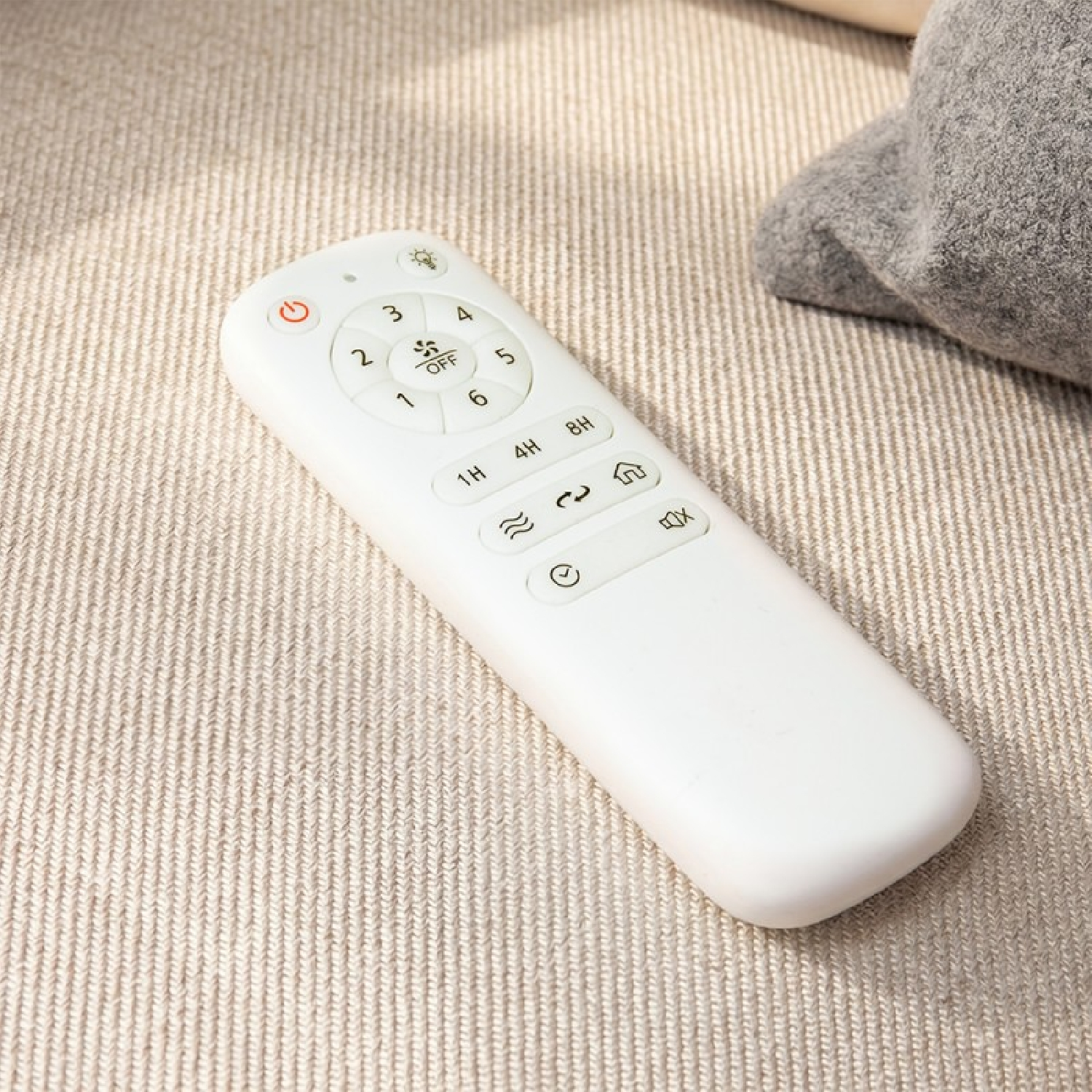 Un control remoto blanco con botones numerados descansa sobre una superficie de tela beige texturizada cerca de un cojín gris.