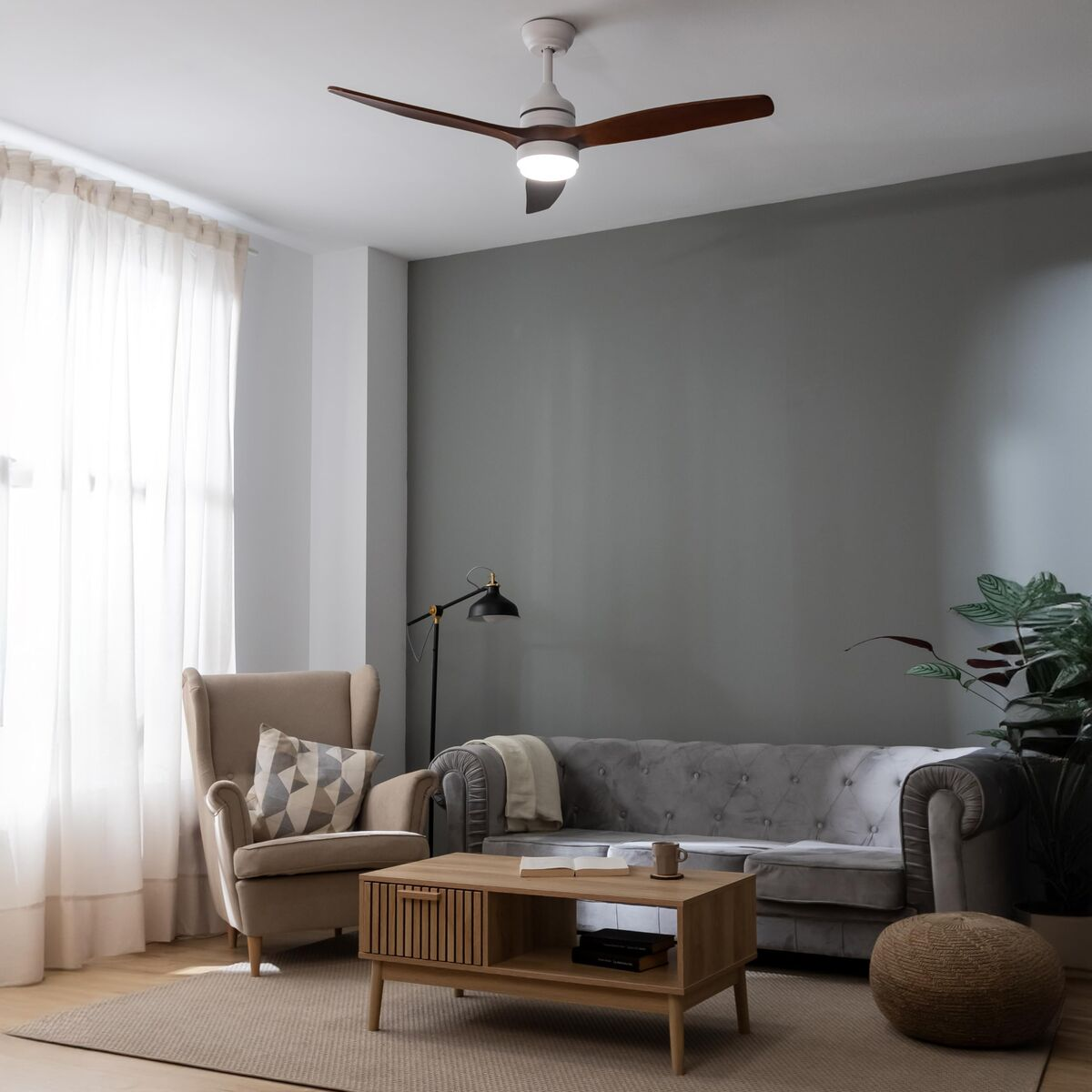 Gemütliches Wohnzimmer mit grauem Sofa, Sessel, Holztisch, Deckenventilator und großem Fenster mit weißen Vorhängen.
