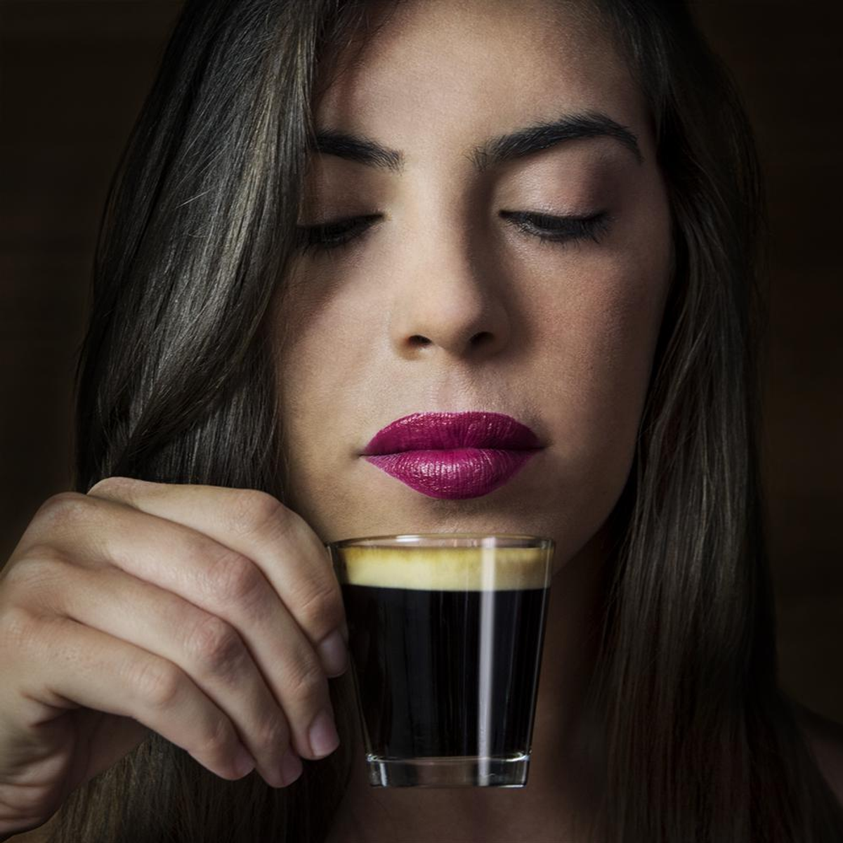Una donna con capelli scuri e rossetto rosso tiene un piccolo bicchiere di espresso davanti al suo viso.