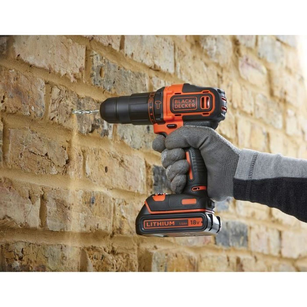 Una mano guantata fora un muro di mattoni con un trapano a batteria nero e arancione.