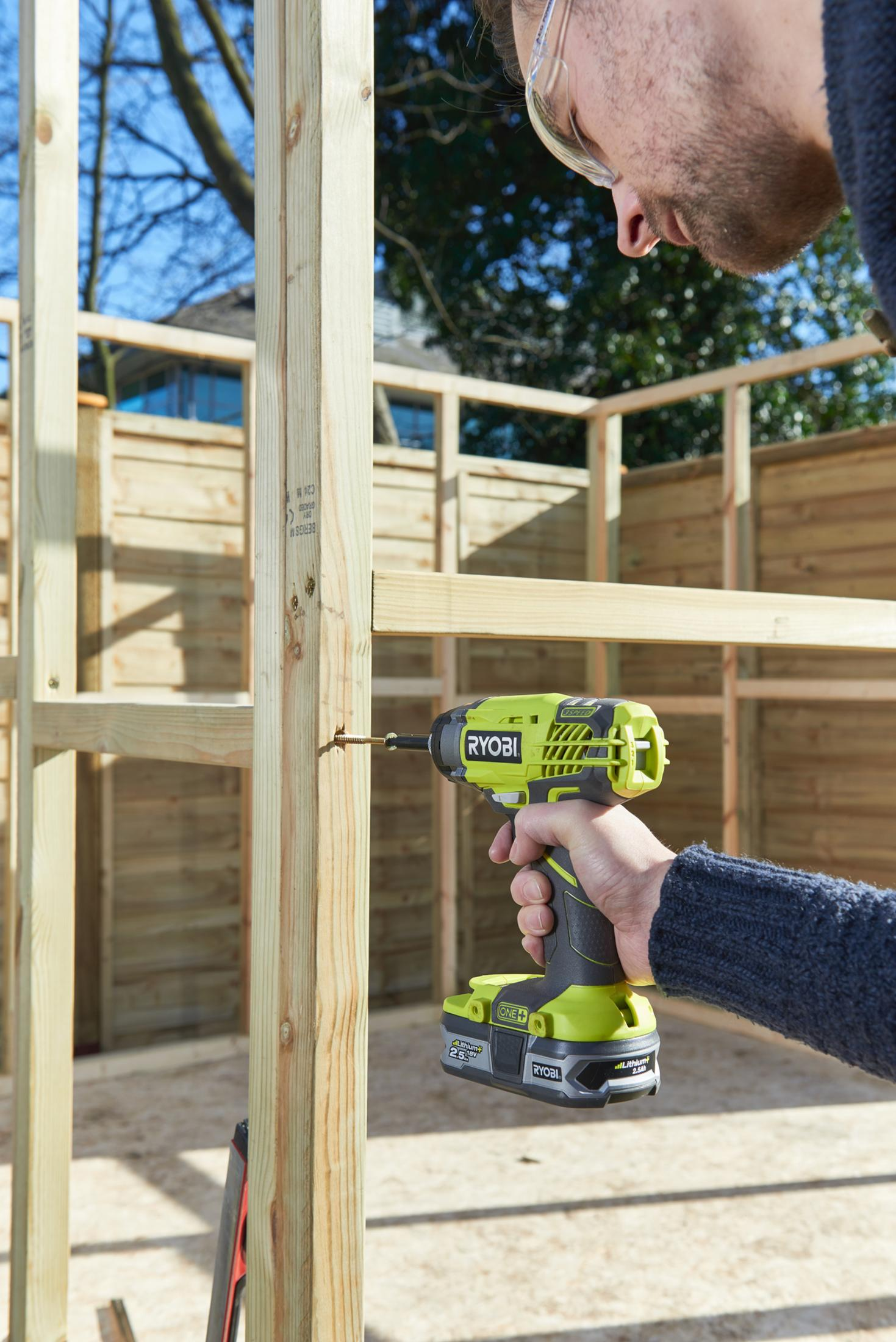Un artigiano avvita travi di legno per una struttura da esterno usando un trapano a batteria verde.