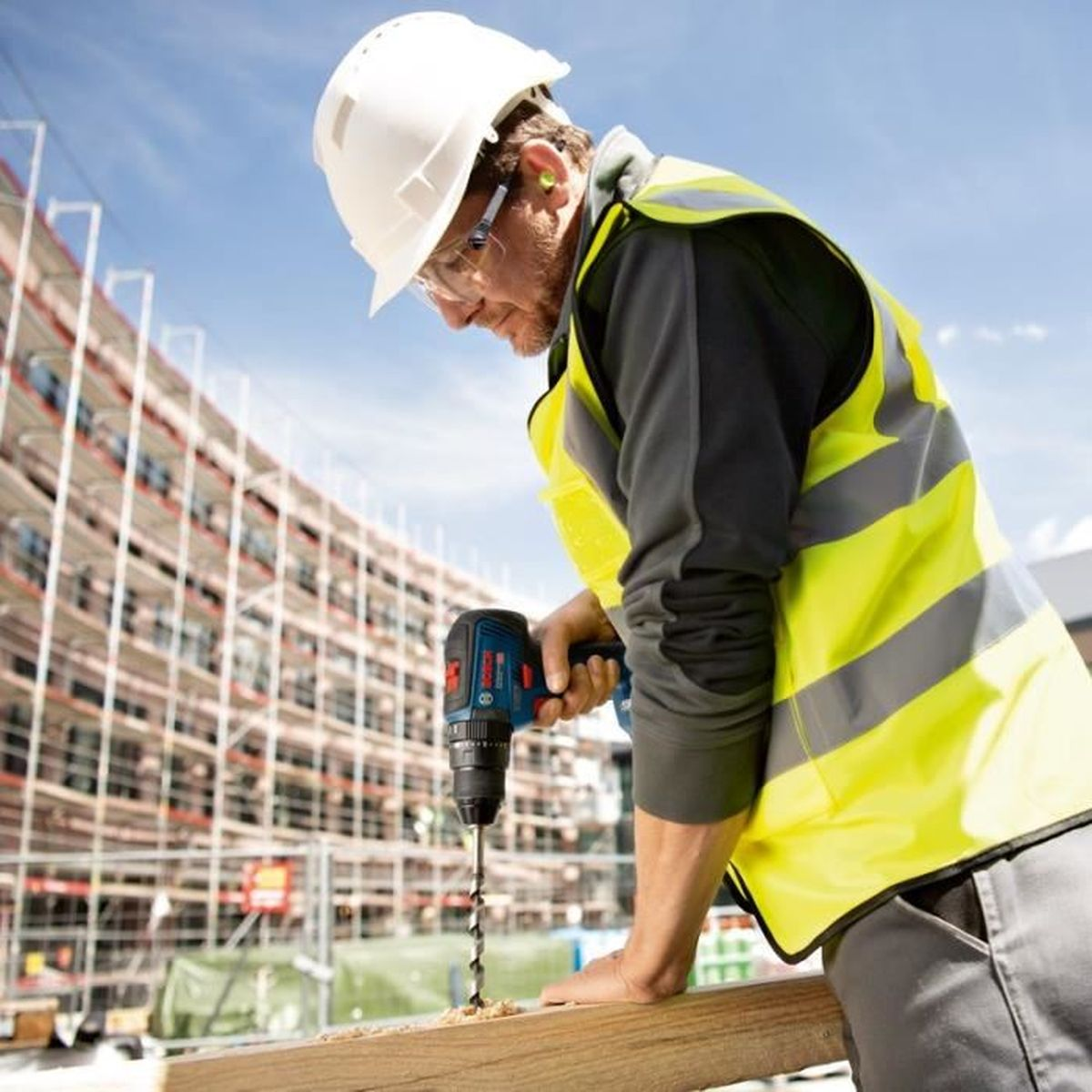 Bauarbeiter mit weißem Helm und gelber Weste bohrt in Holz vor einer Baustelle.